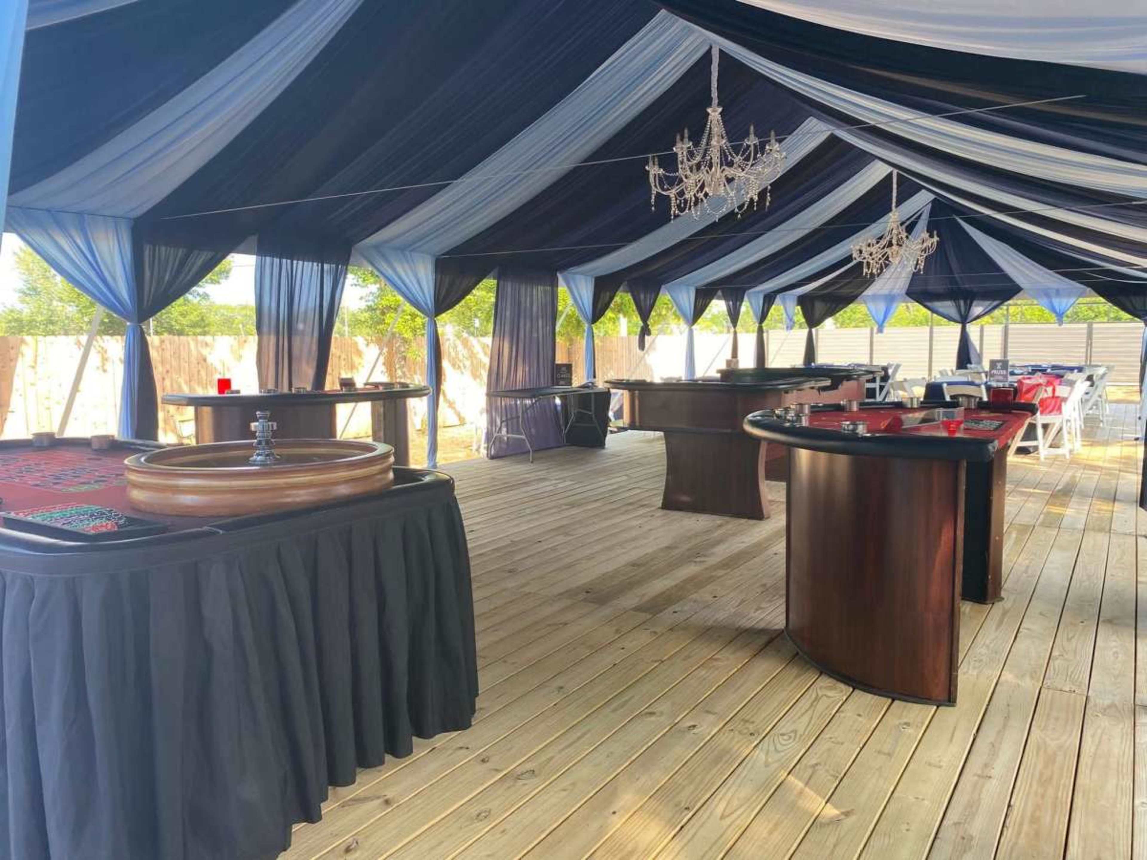 The image shows a large outdoor tent decorated with blue and black drapery, featuring casino tables arranged on a wooden deck.