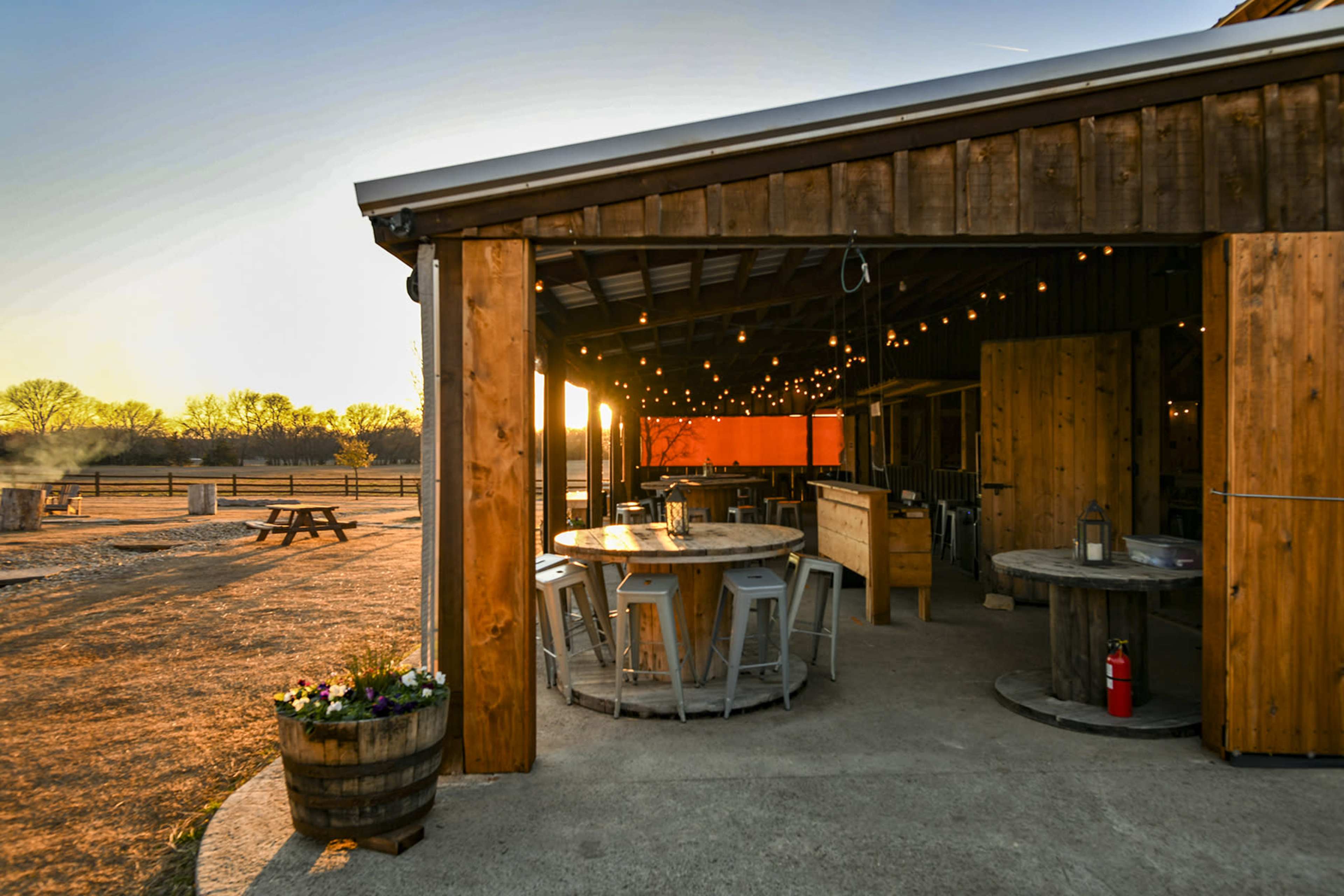 The image shows an outdoor gathering area with wooden structures, round tables, and string lights, illuminated by a sunset.