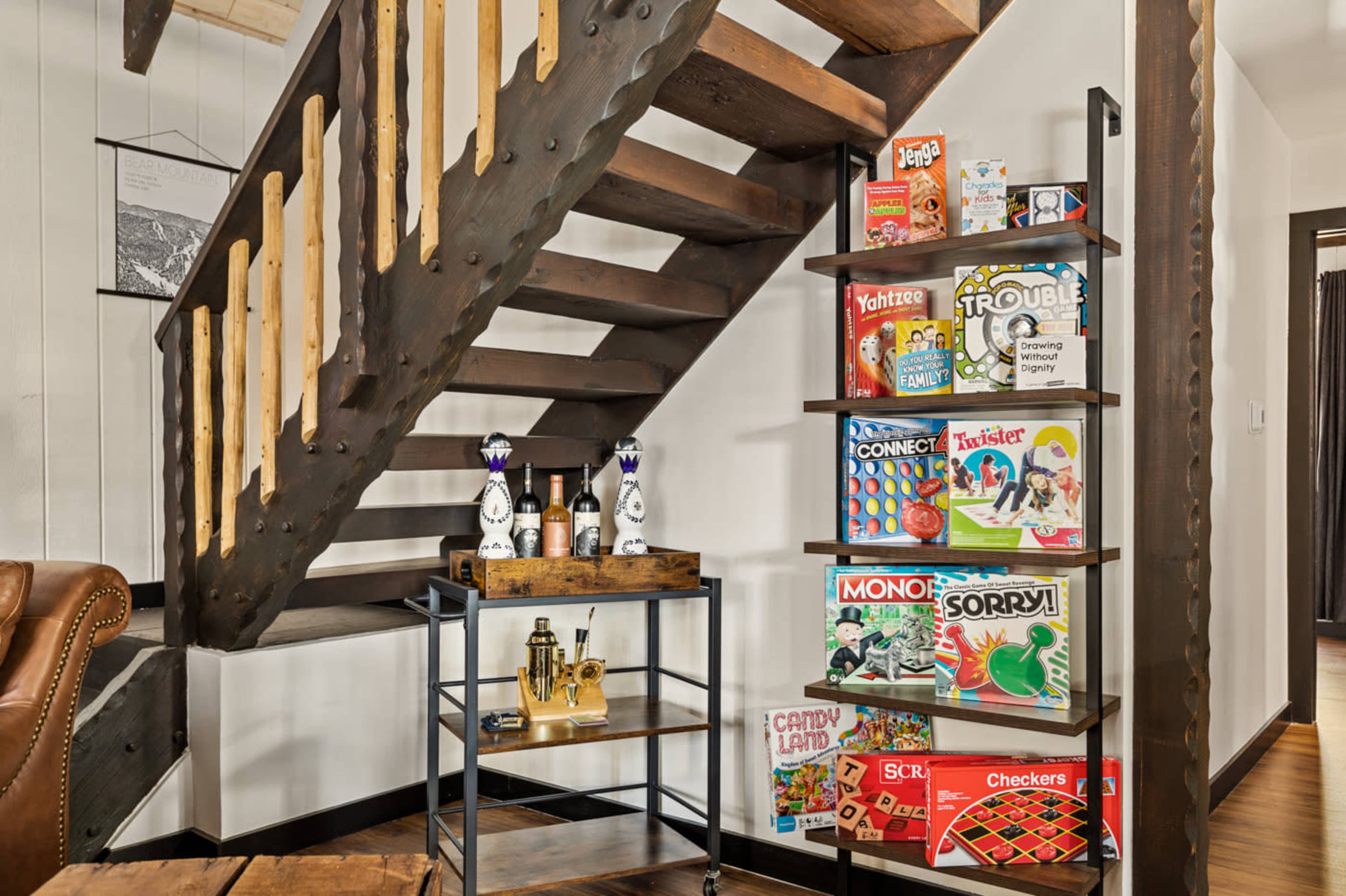 The image shows a wooden staircase next to a shelving unit filled with board games and a cart with bottles and barware.