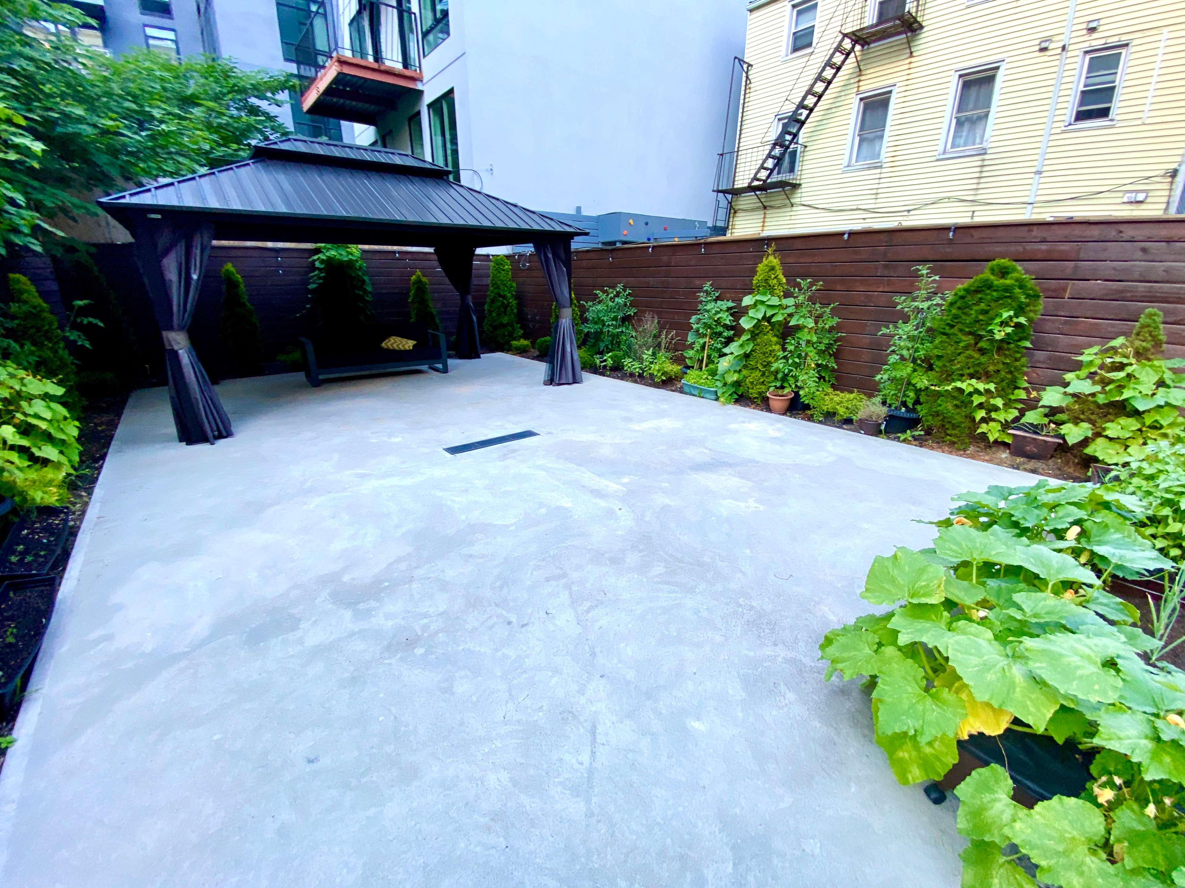 A concrete patio features a gazebo surrounded by greenery, including potted plants and garden beds.