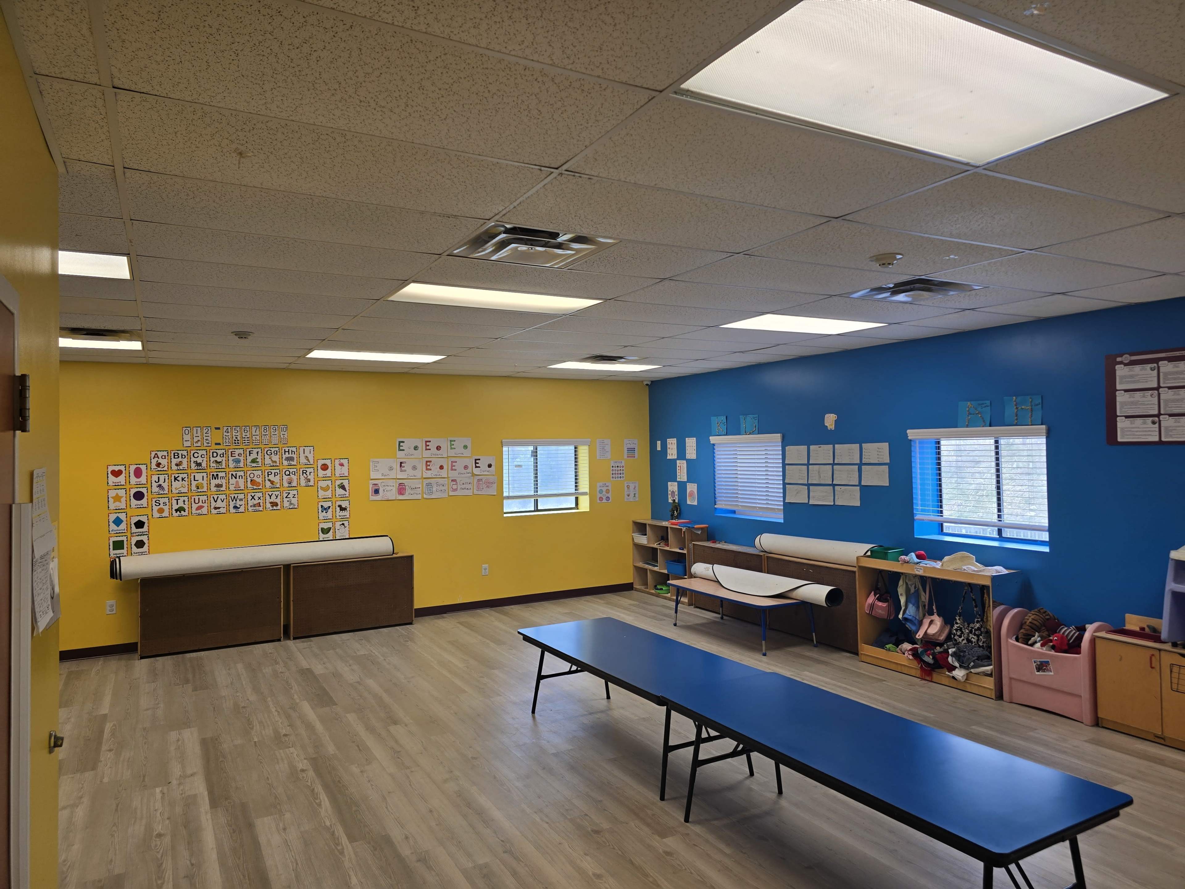 The image shows a brightly colored classroom with yellow and blue walls, a table in the center, and various educational materials displayed on the walls and shelves.