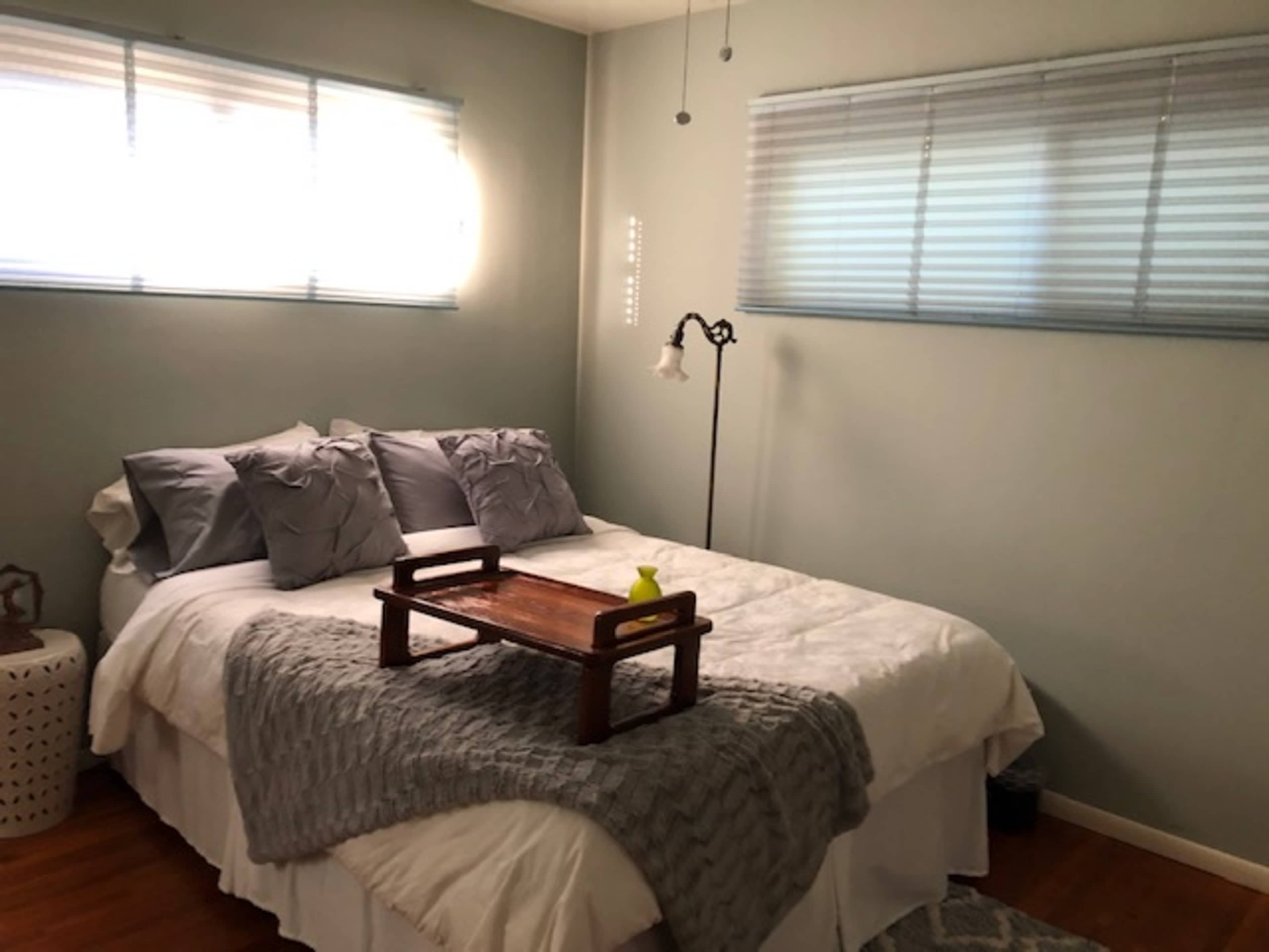 A neatly arranged bedroom with a bed covered in white linens, two gray pillows, and a wooden tray on top, situated near two windows with blinds.
