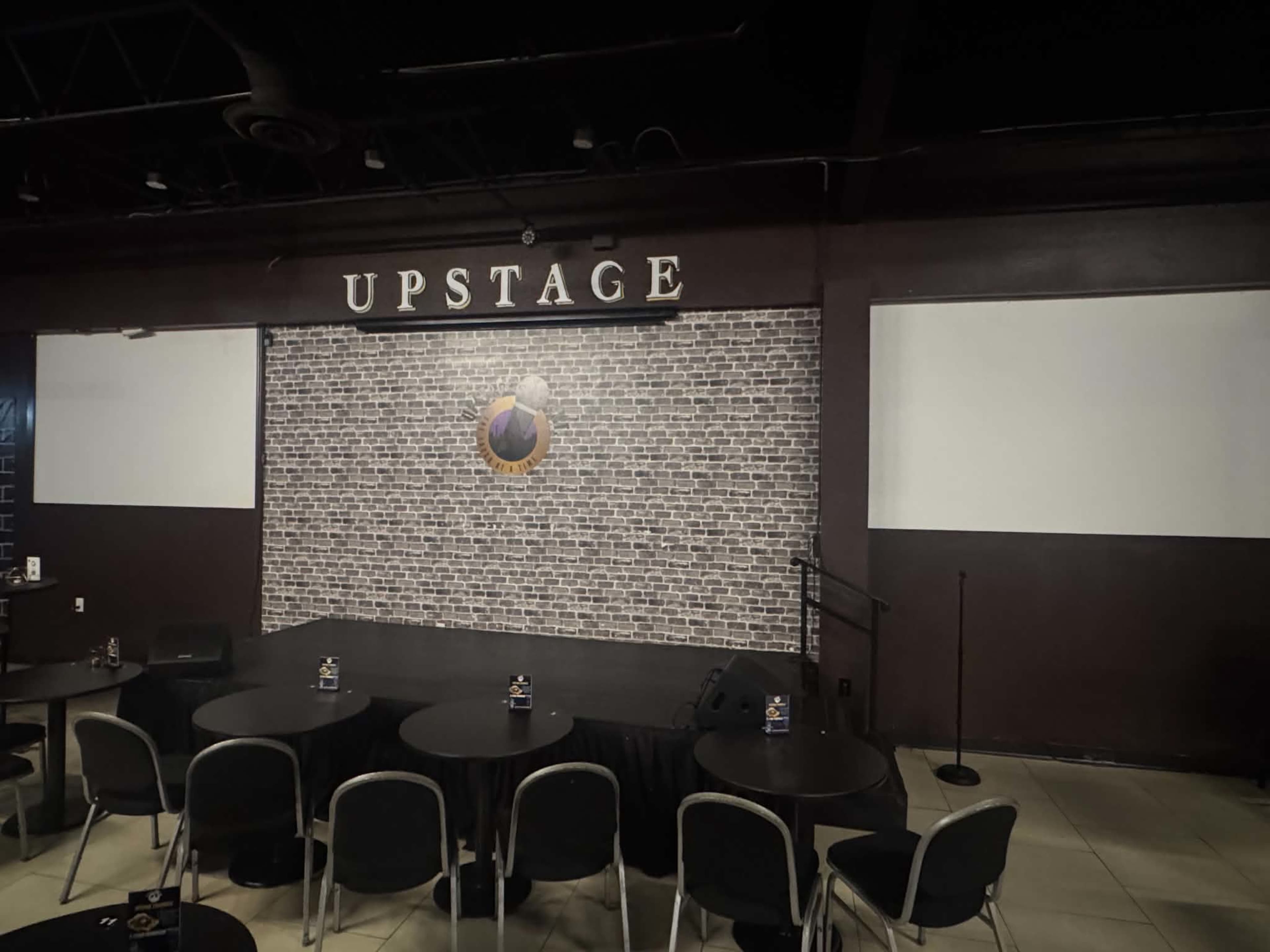 The image shows a stage area with a sign reading "UPSTAGE" above a brick-patterned backdrop, flanked by two empty white projection screens on either side.