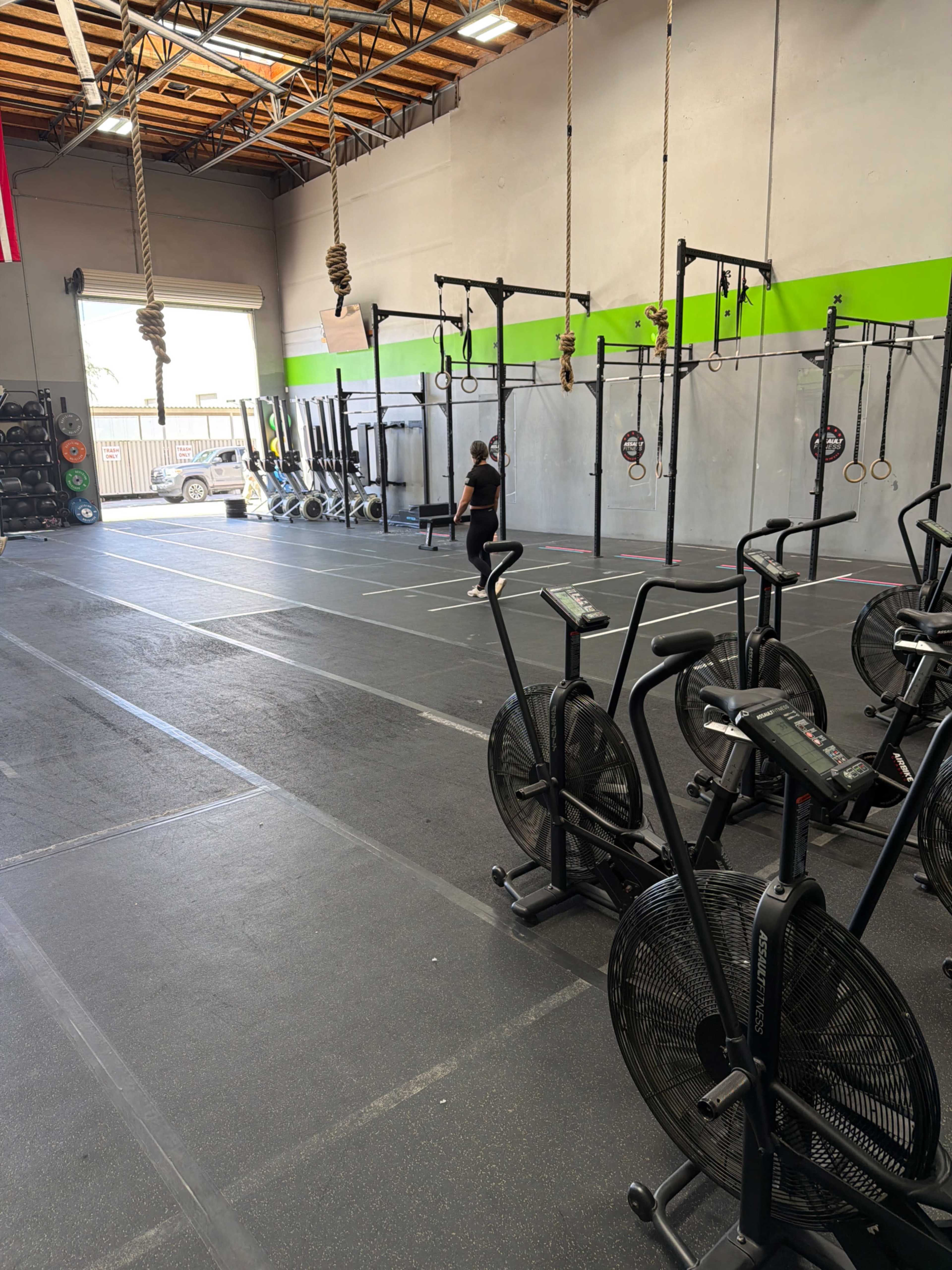 The image shows an empty gym space with exercise equipment such as air bikes and a training area featuring pull-up bars and other workout stations.