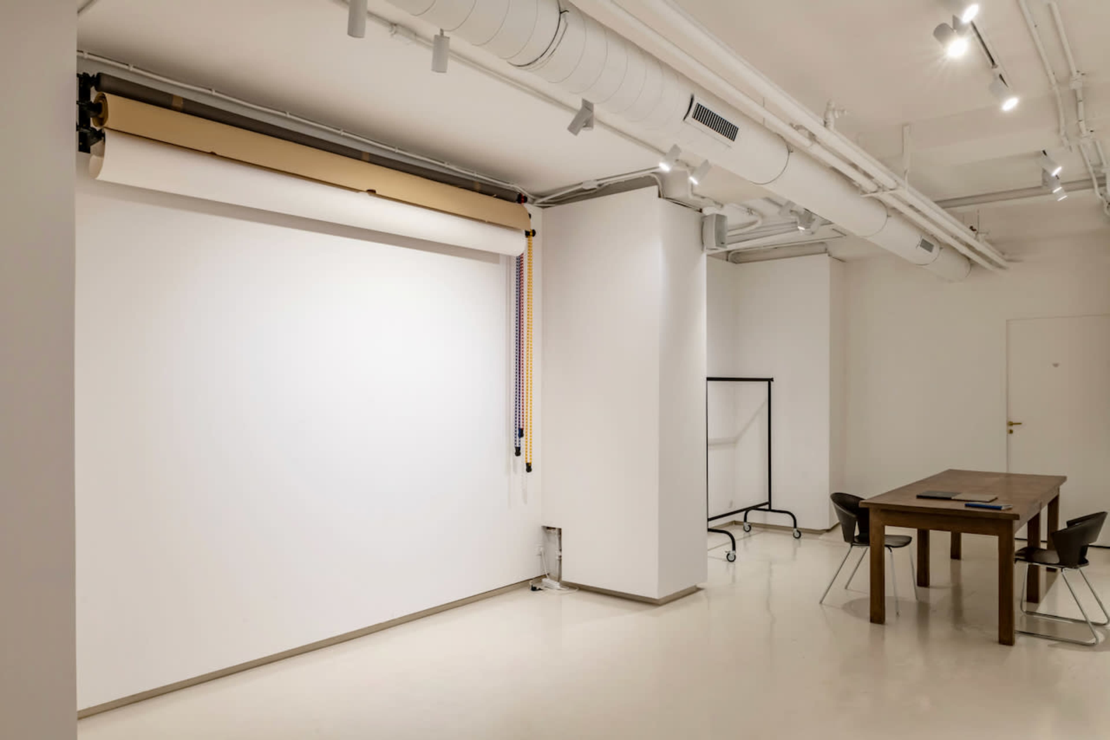 The image shows a minimalist studio space featuring a large blank wall with a pull-down backdrop, a wooden table, and a few chairs.