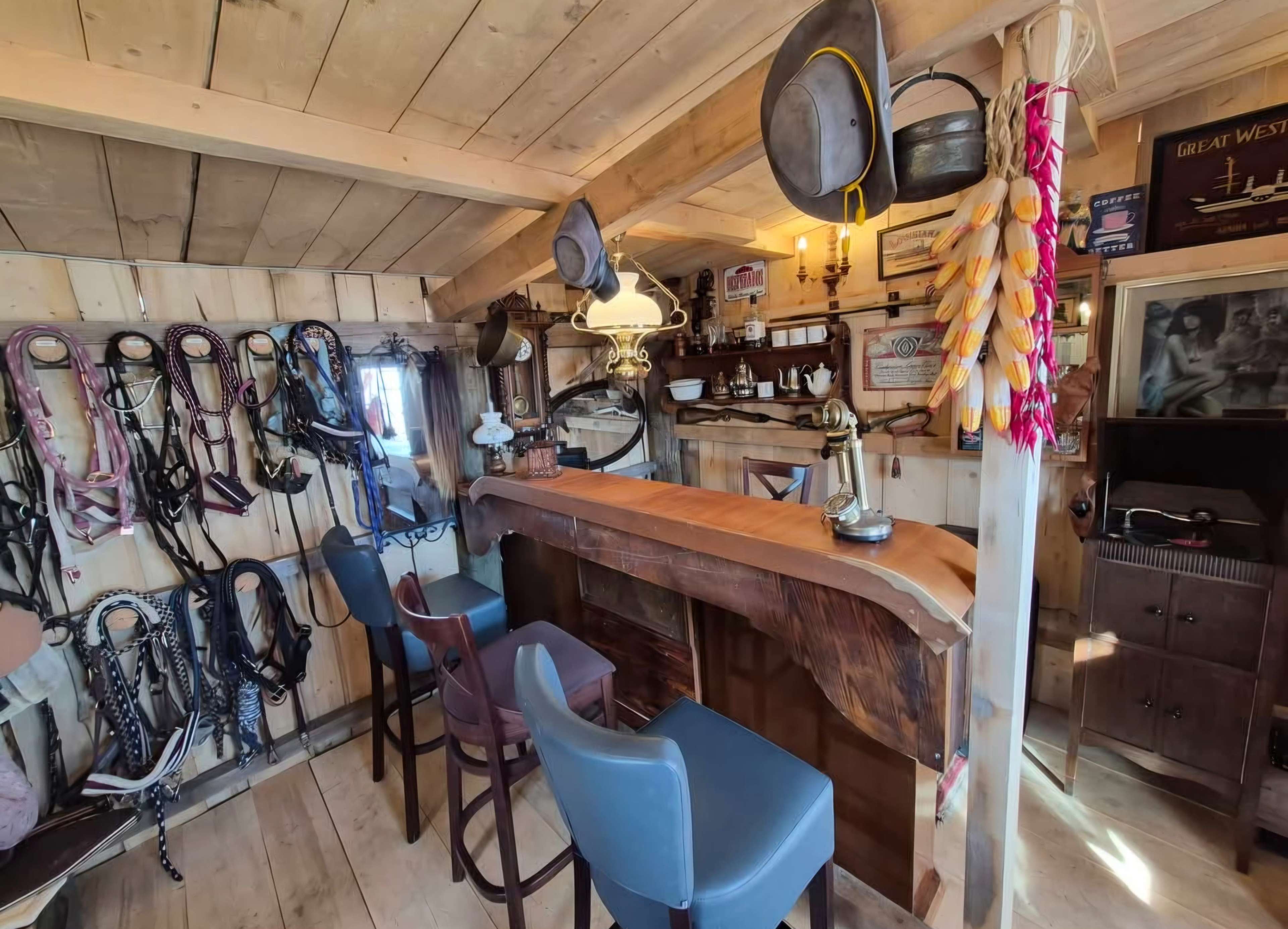 The image shows a rustic interior of a bar with wooden walls, a polished wooden counter, and several bar stools, surrounded by horse tack and western decor.
