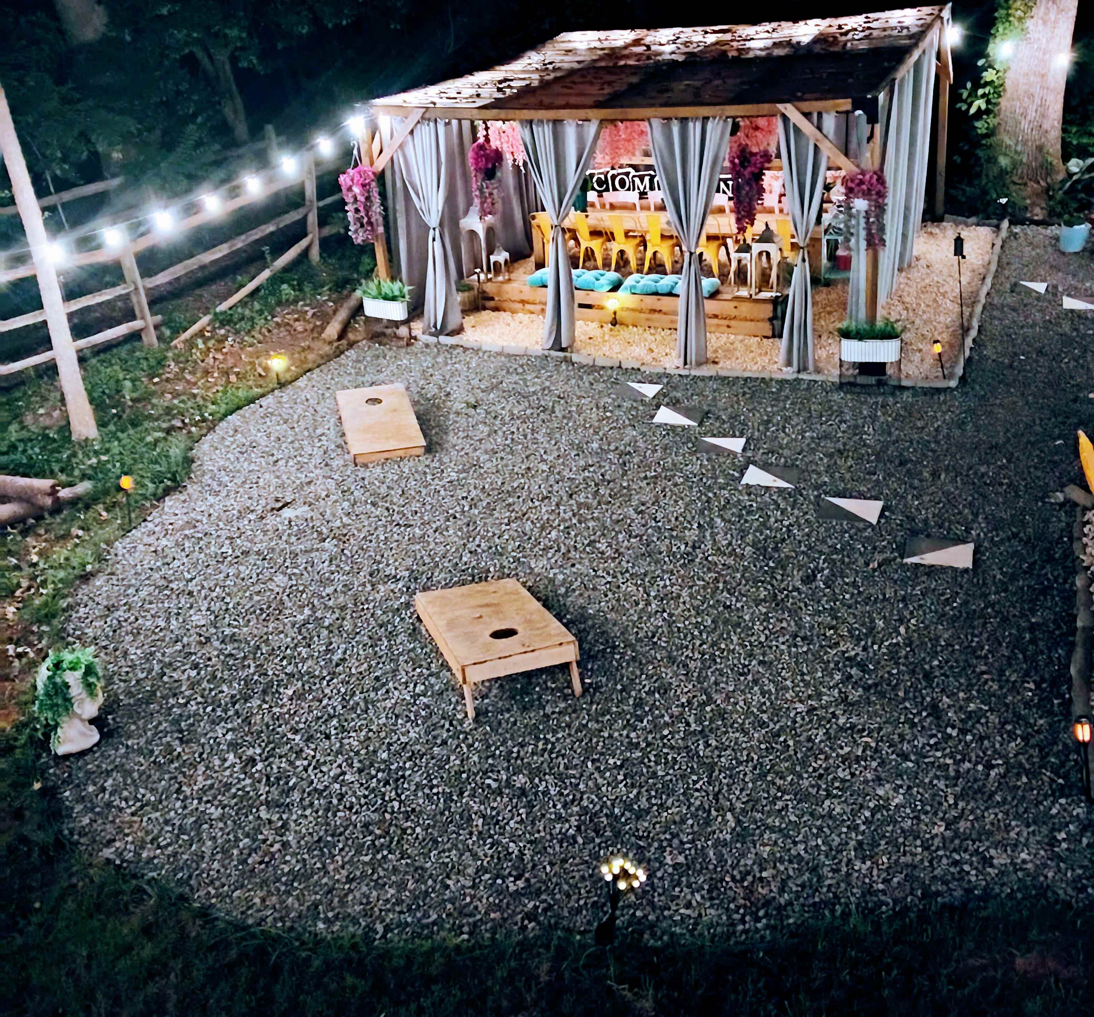 The image shows an outdoor setup featuring a wooden structure draped with curtains, surrounded by gravel pathways and string lights, with several chairs arranged inside.
