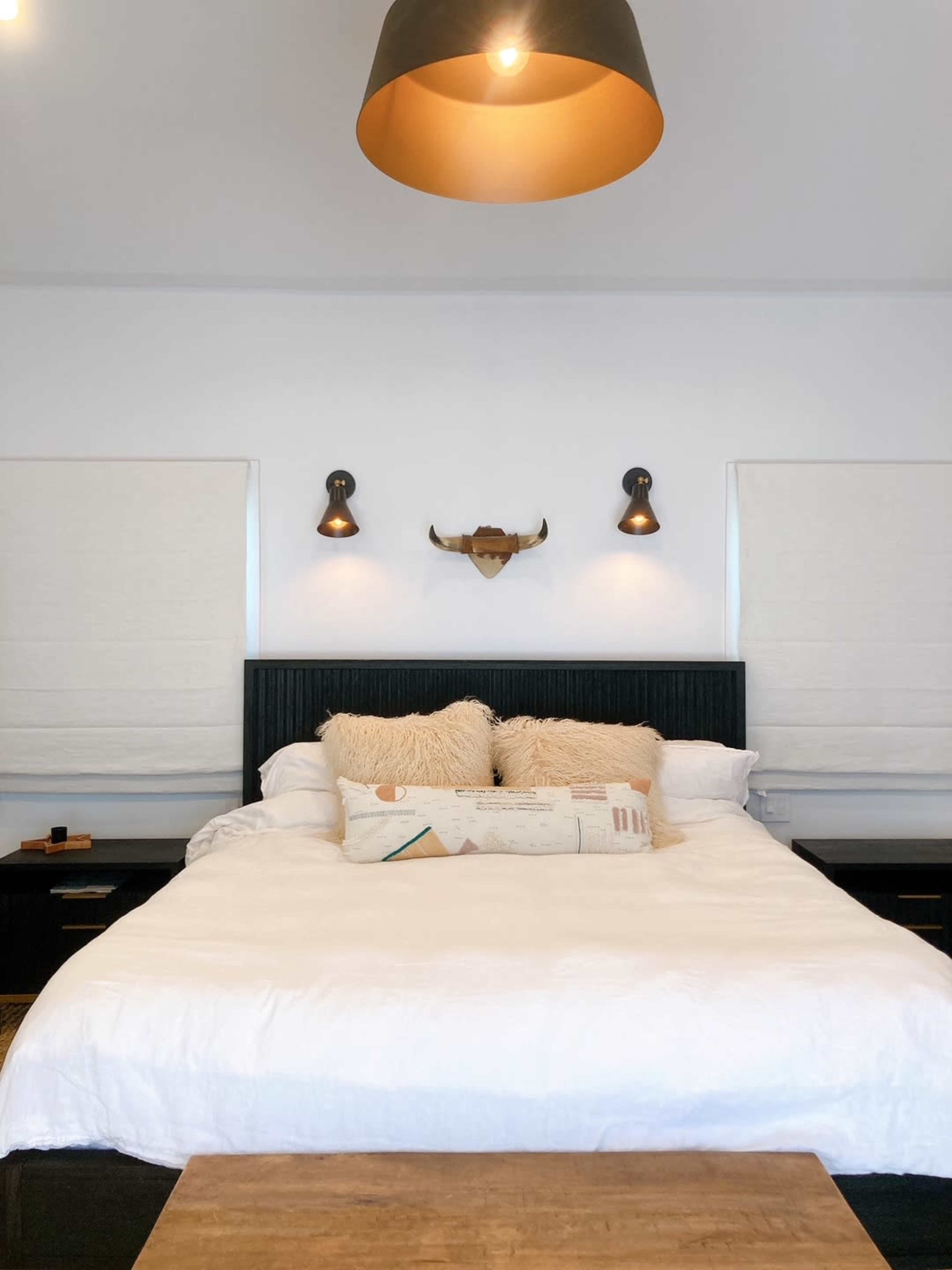 The image shows a neatly arranged bedroom with a large bed, decorative pillows, and mounted wall sconces on either side.
