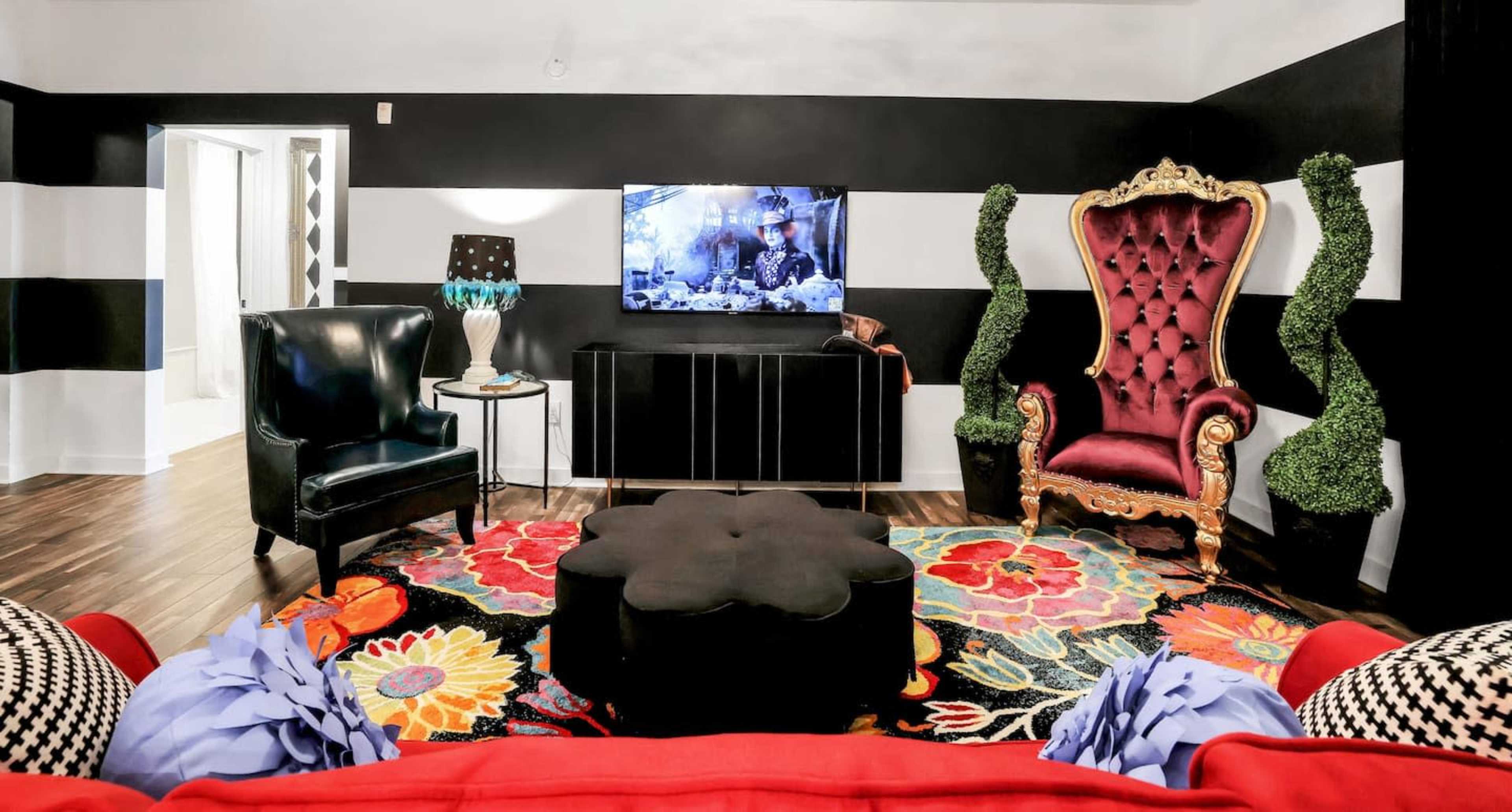 The living room features a large black and red area rug, two distinct chairs, a television mounted on the wall, and decorative green plant structures.