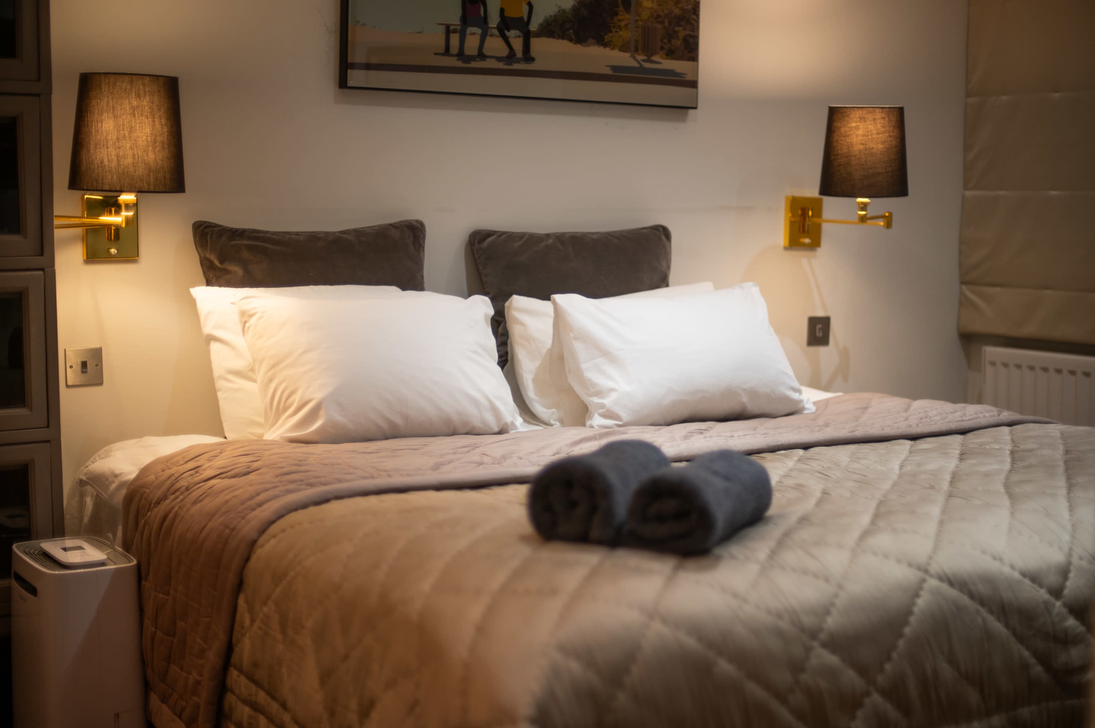 The image shows a neatly made bed with two white pillows and a beige quilt, flanked by two wall-mounted lamps in a softly lit bedroom.