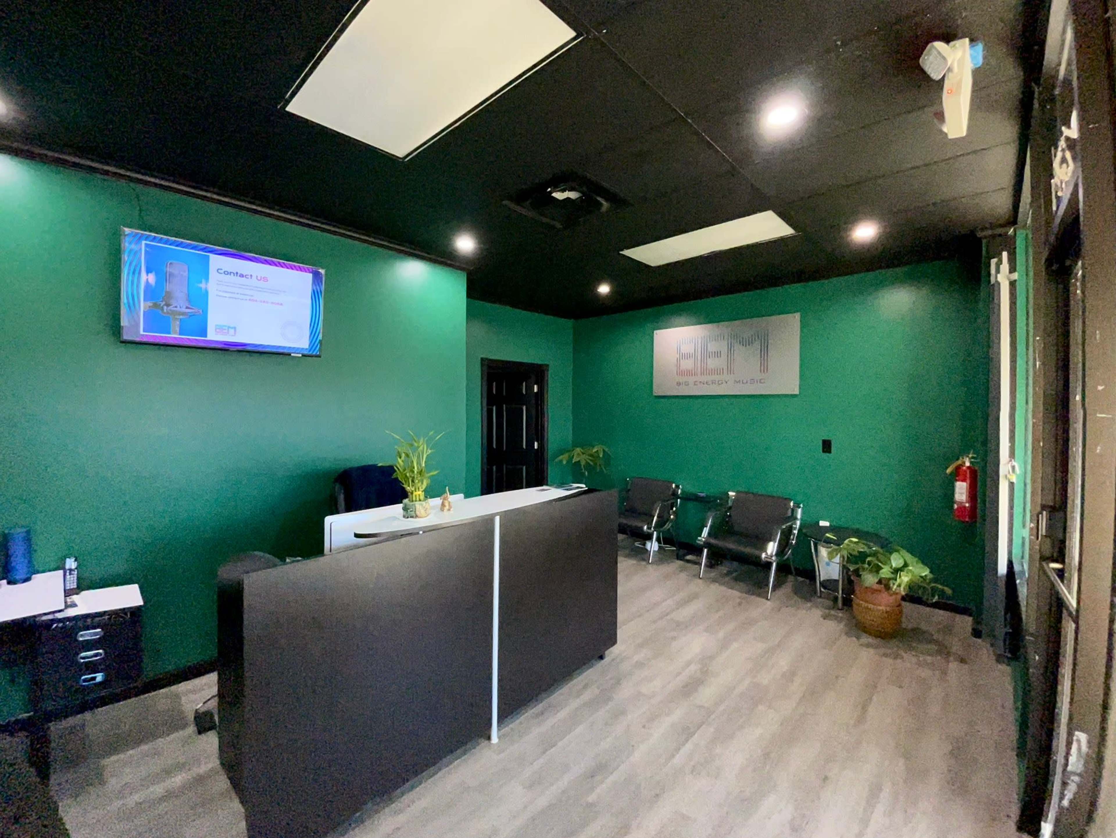 The image shows a small reception area with a dark green wall, a front desk, a television displaying information, and several chairs arranged for visitors.