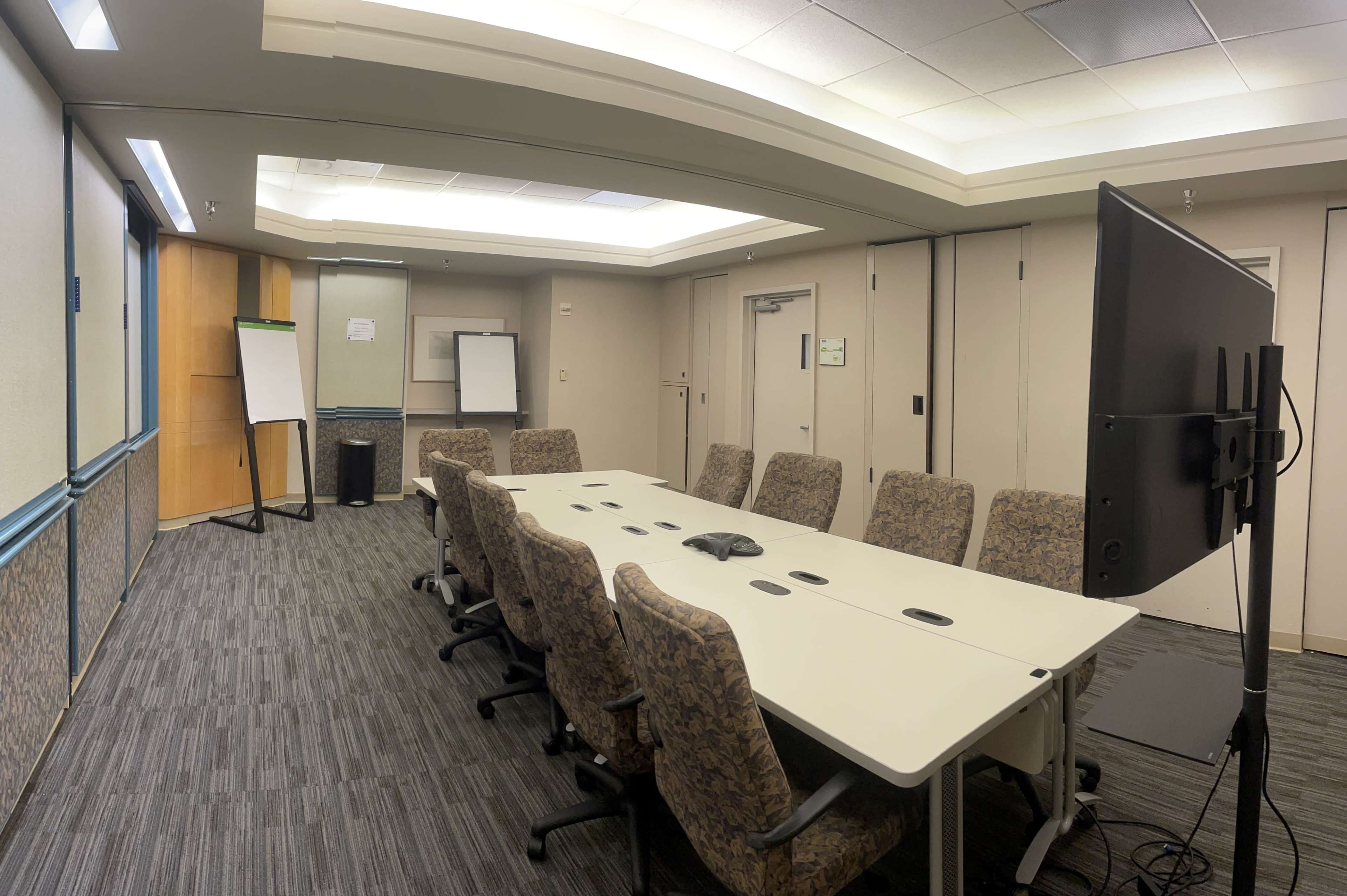 A conference room with a long table surrounded by eight chairs, featuring a large screen and a whiteboard.