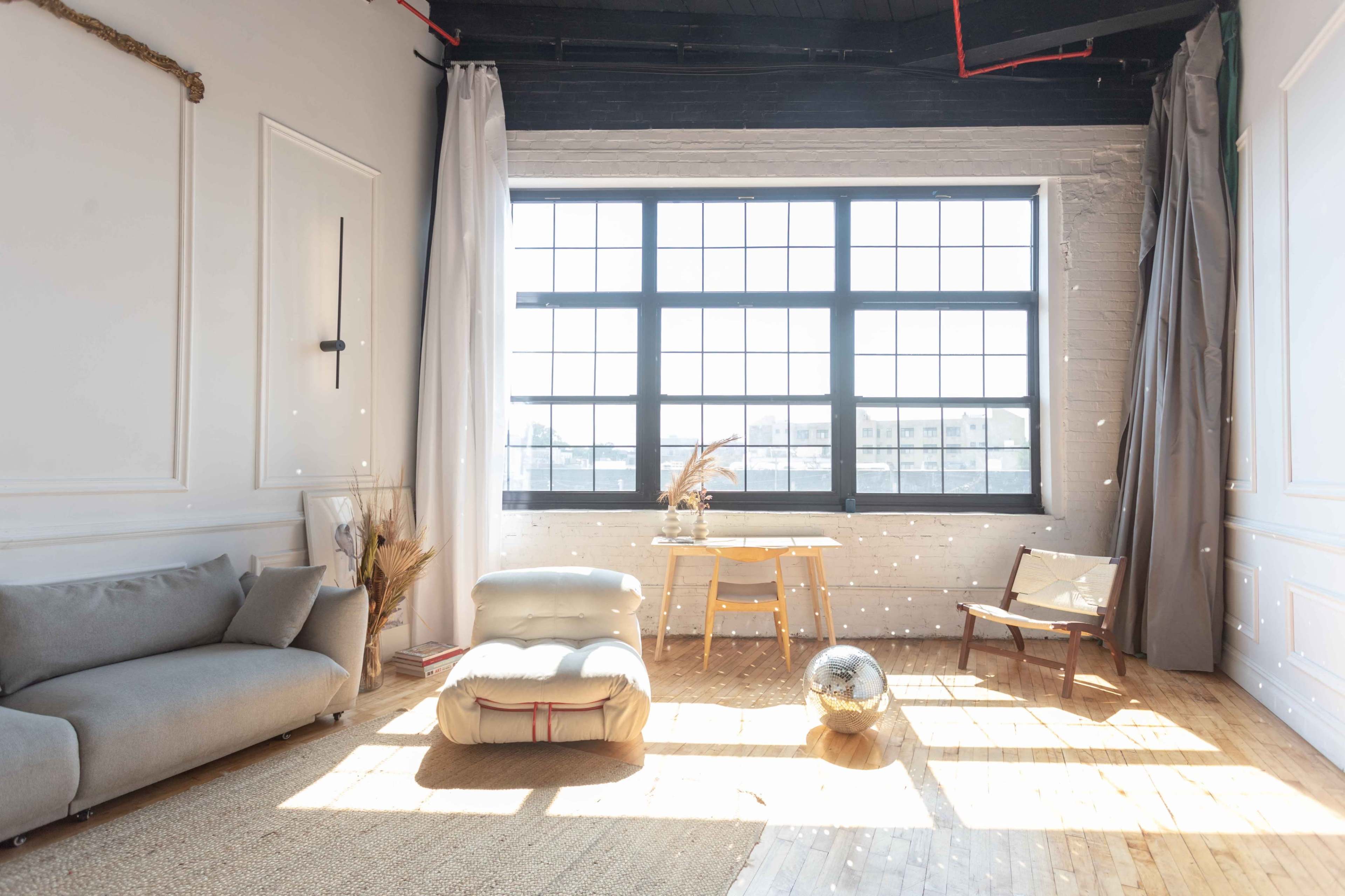 A spacious, sunlit room features a gray sofa, a low white cushion, a wooden table, and a decorative globe, with large windows allowing natural light to fill the space.