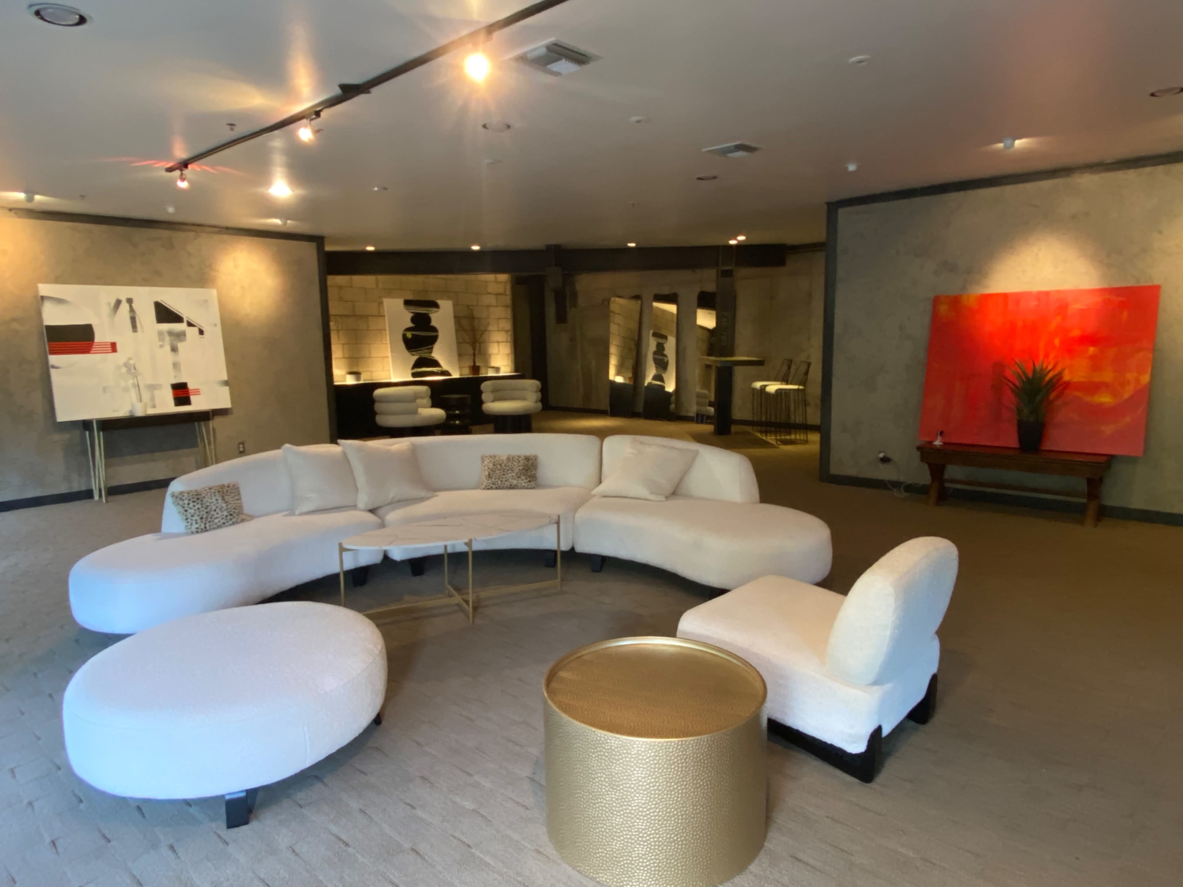 The room features a large, curved white sectional sofa arranged around a circular coffee table, with abstract art displayed on the walls and a wooden table in the background.