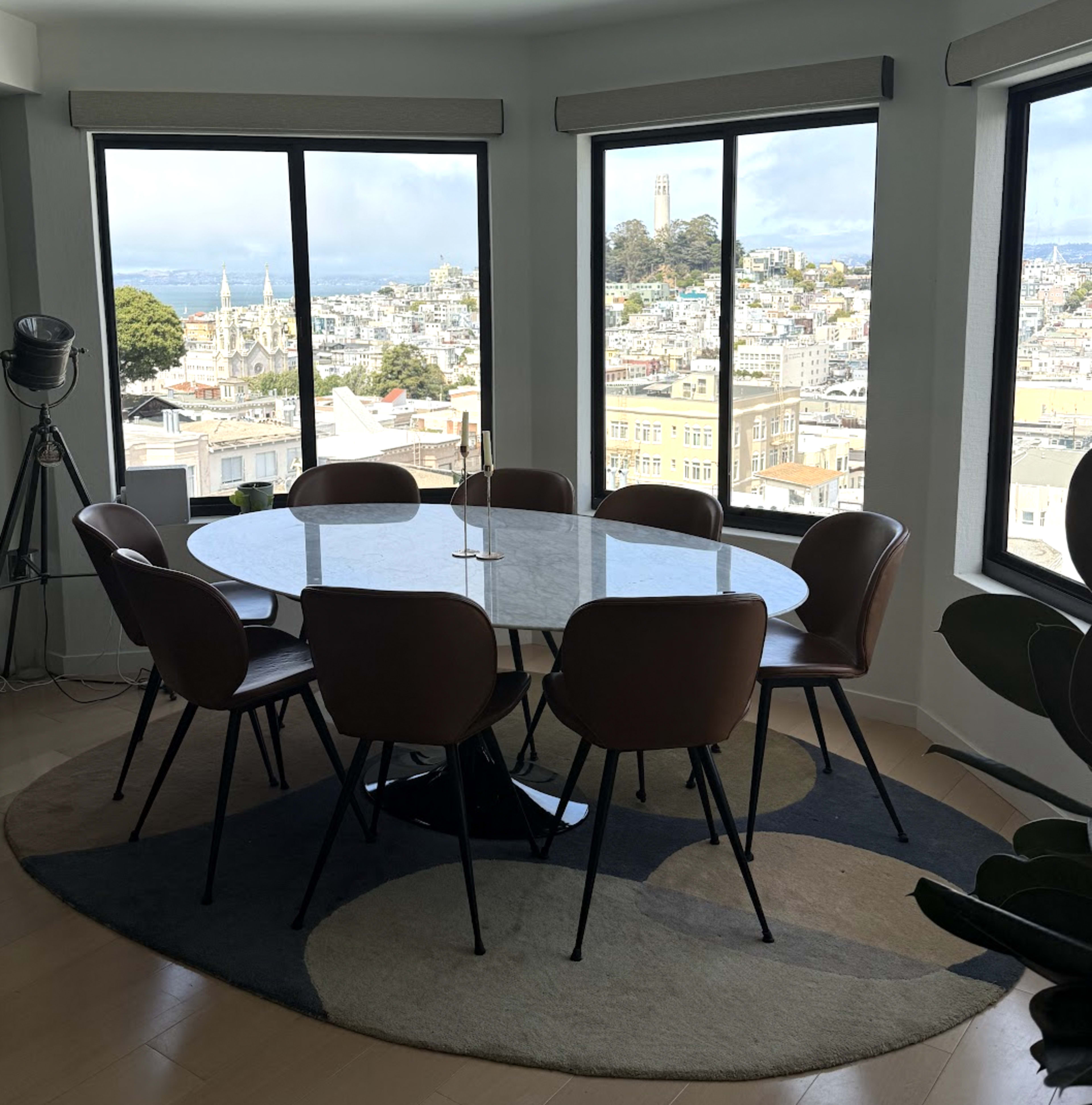 A modern dining area features a round marble table surrounded by eight brown chairs, with large windows showcasing a cityscape view.