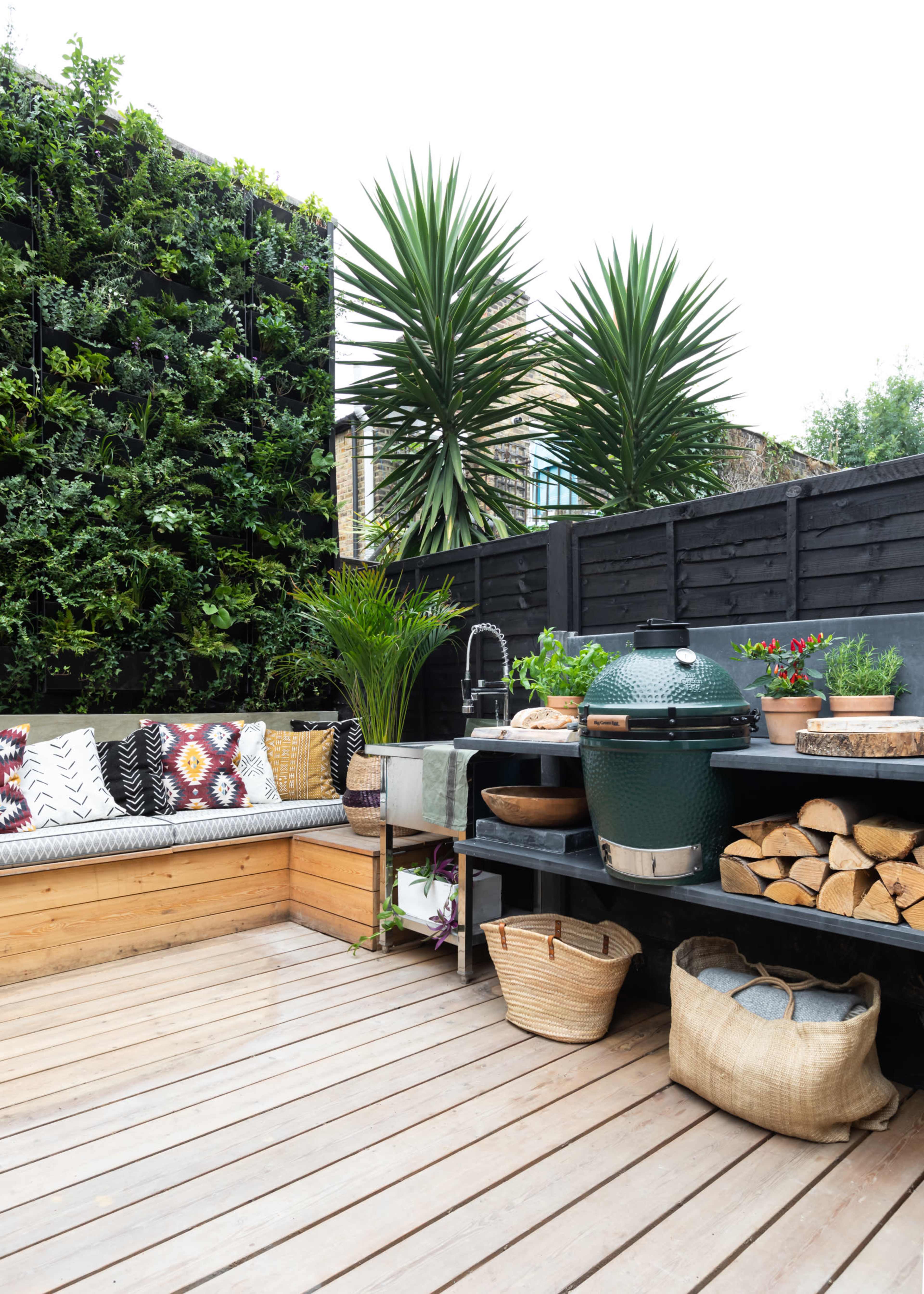 A patio area featuring a corner seating arrangement with decorative cushions, a large green egg grill, and neatly stacked firewood beside a variety of potted plants.