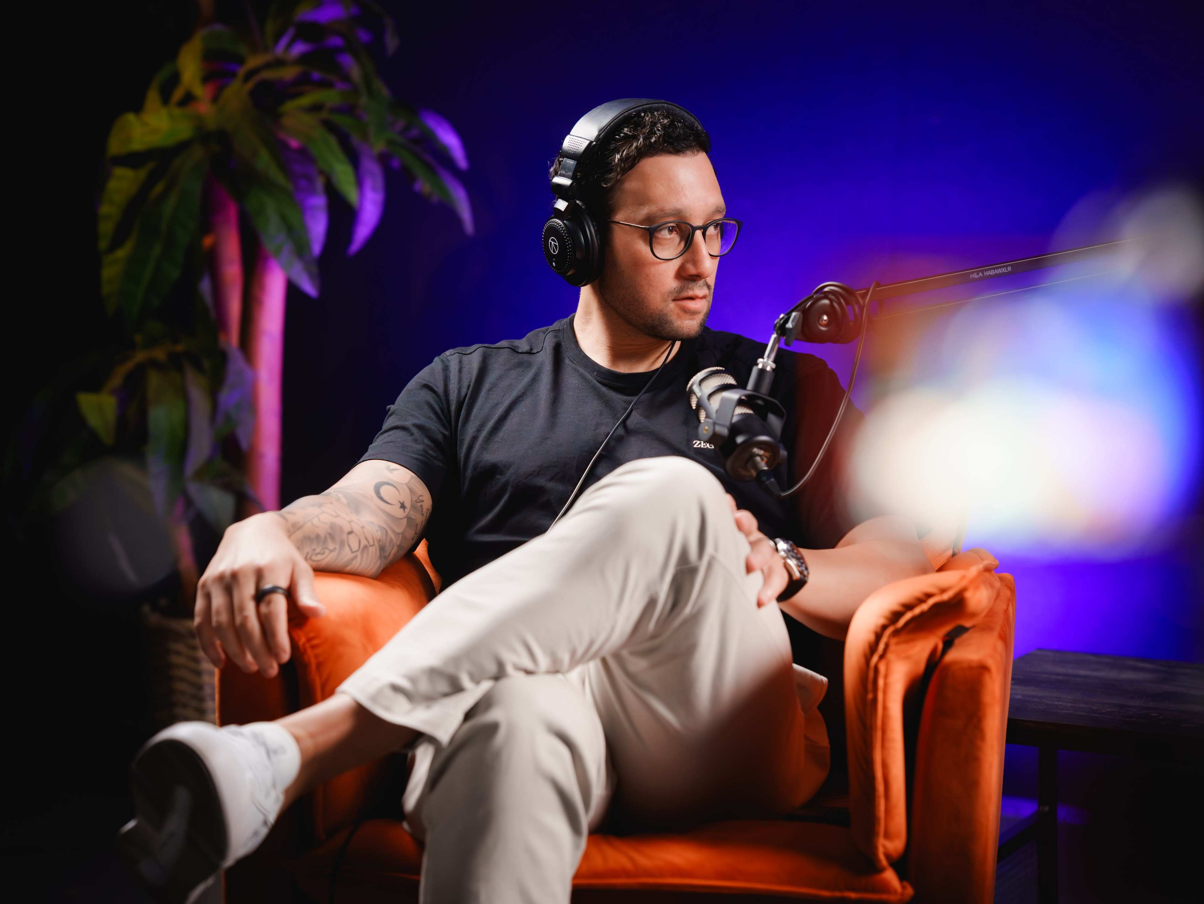 A man with glasses sits on an orange chair, wearing headphones and a black shirt, in front of a microphone with a purple backdrop and a plant nearby.