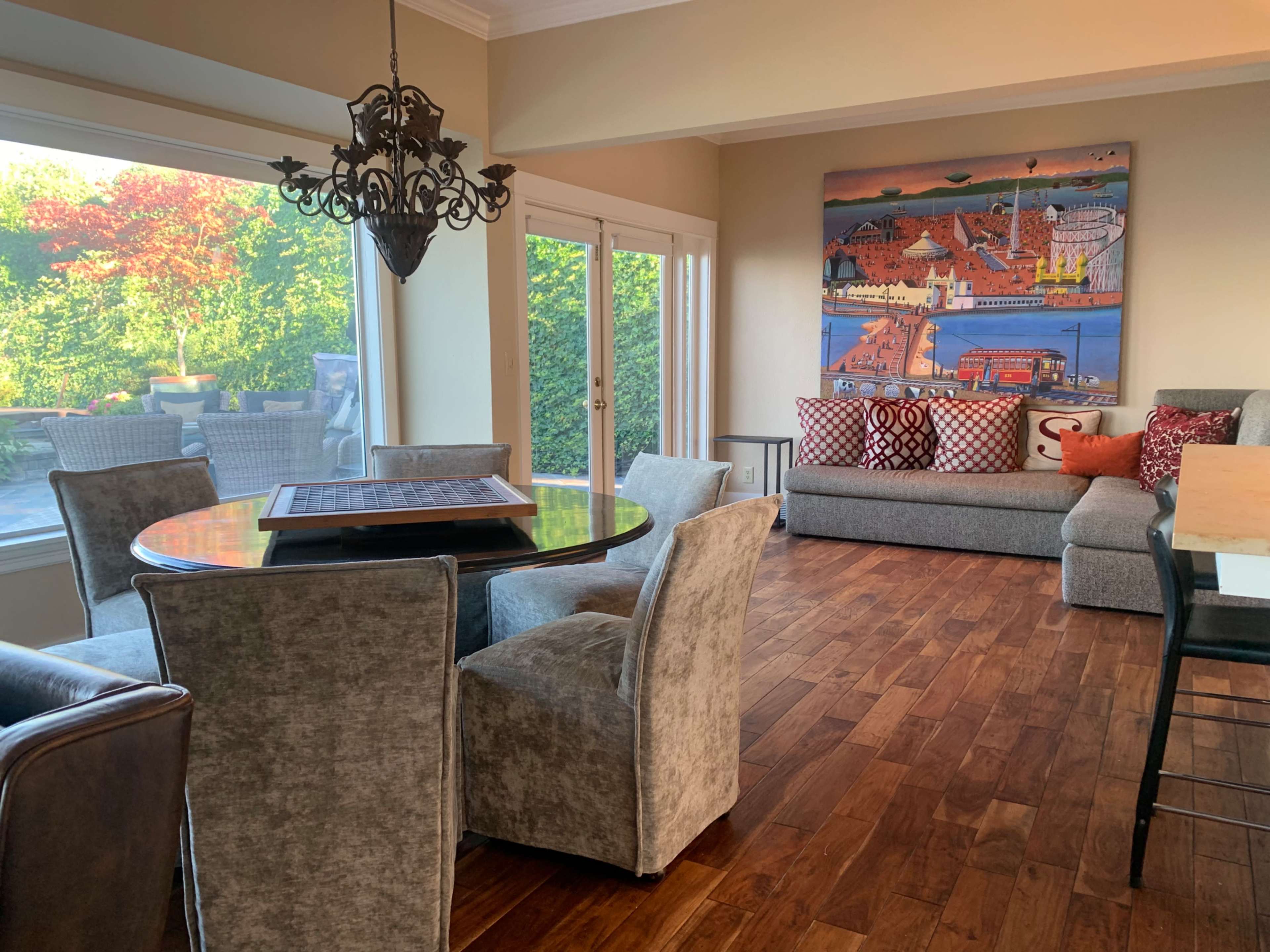 A spacious dining area with a round table, upholstered chairs, and a large artwork on the wall, beside a window that overlooks a greenery-filled outdoor space.