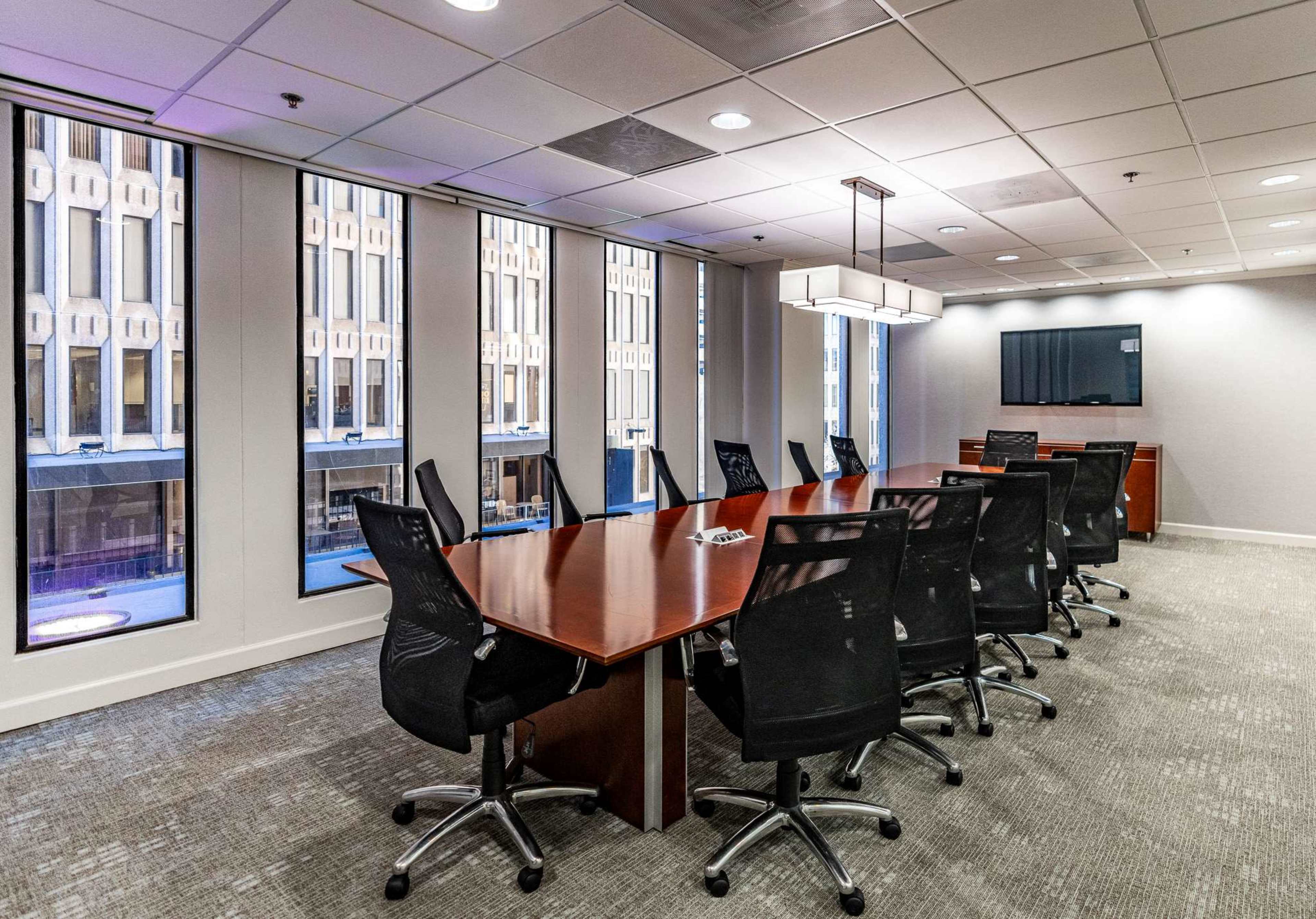 A large, rectangular conference table surrounded by black office chairs is located in a well-lit room with windows displaying an urban view.