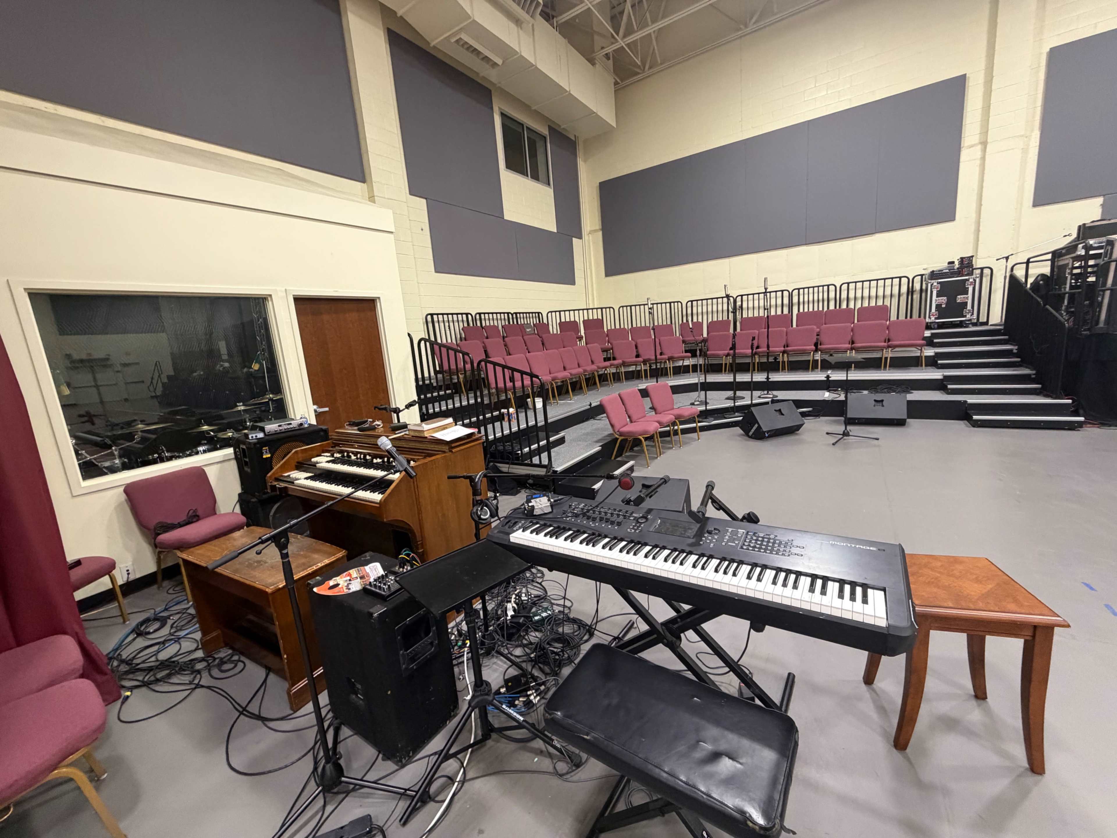 The image shows a music performance space with a keyboard, an organ, and scattered cables in the foreground, along with seating arranged in rows behind.