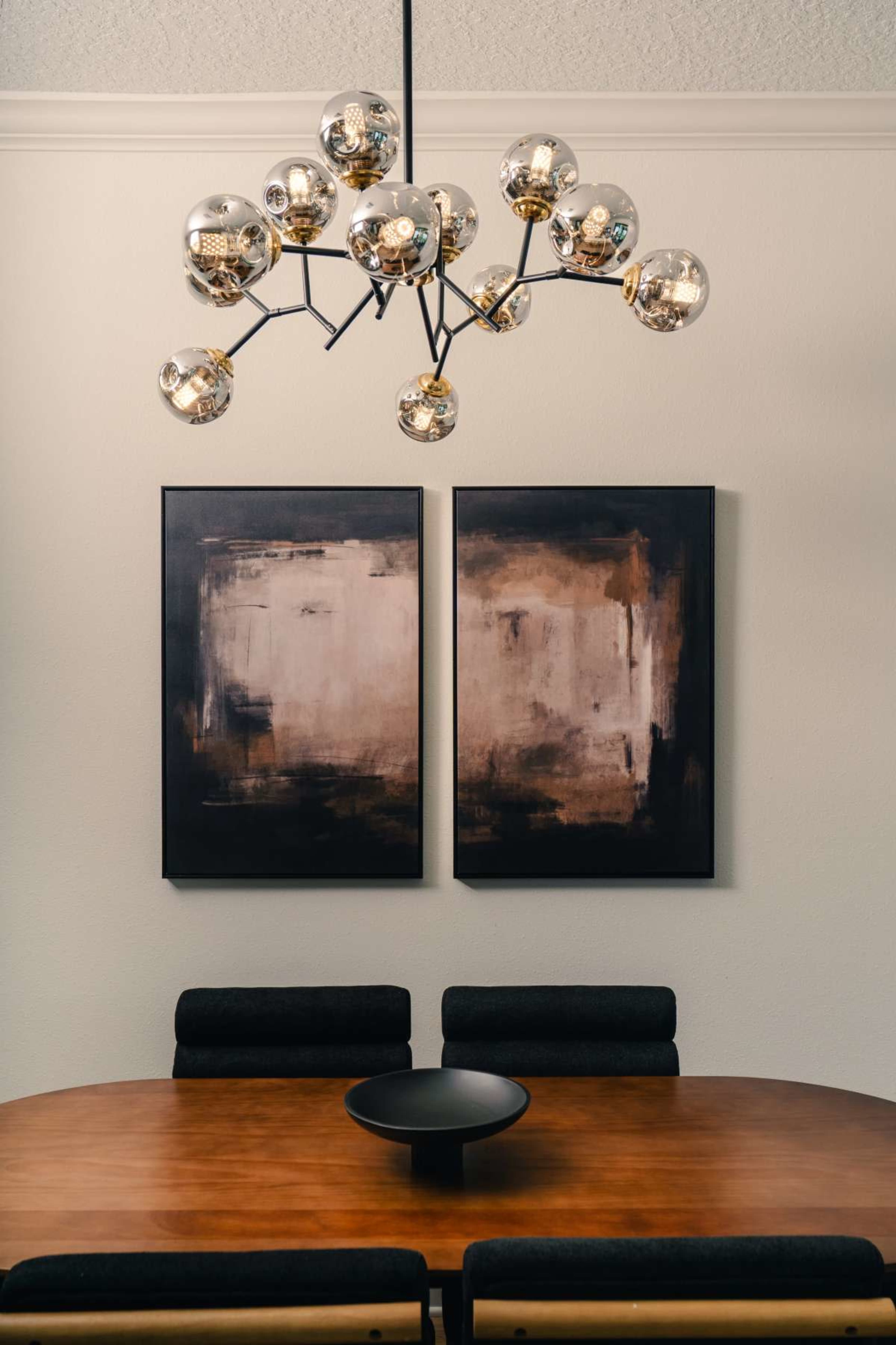 A round wooden dining table is set beneath a modern chandelier, with two abstract paintings hung on the wall above.