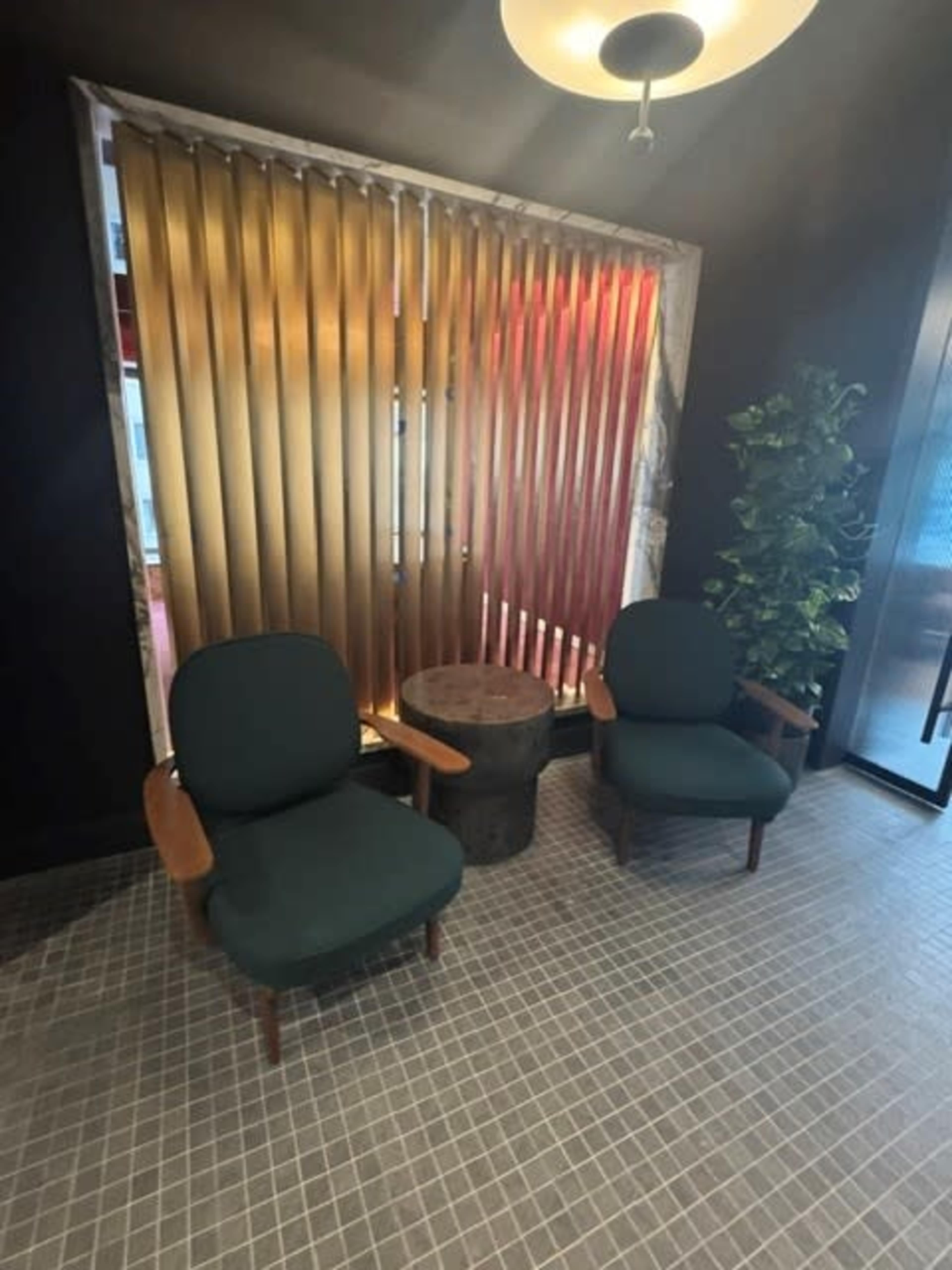 The image shows a cozy corner with two green chairs, a wooden side table, and a window adorned with vertical blinds, against a dark wall.