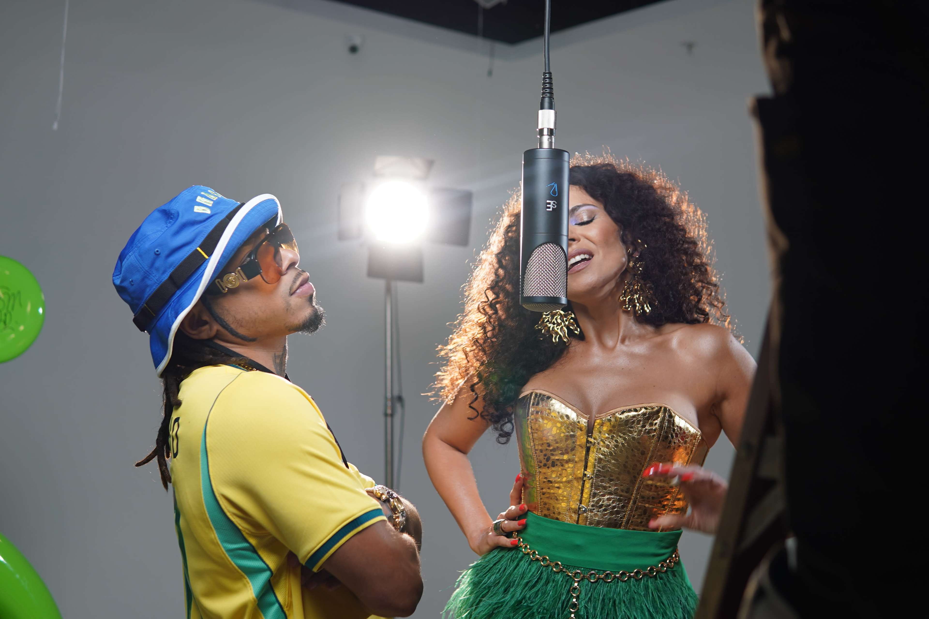 A man in a colorful shirt and sunglasses stands beside a woman in a gold top and green skirt as they perform in front of a microphone in a studio setting.