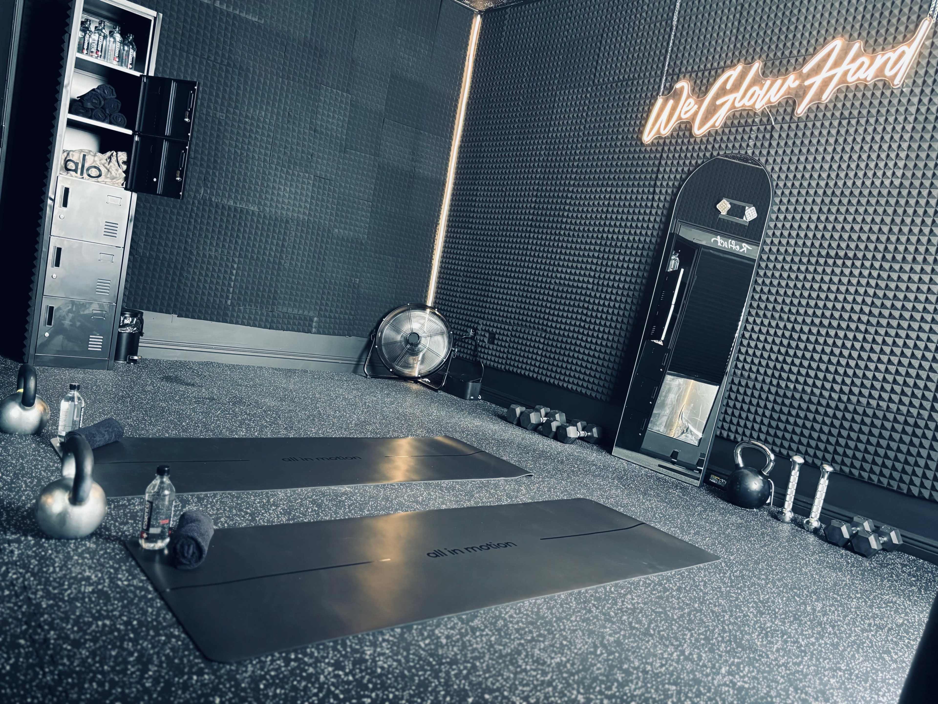 A modern gym interior with two exercise mats, kettlebells, water bottles, and a fan against a textured wall featuring neon signage.