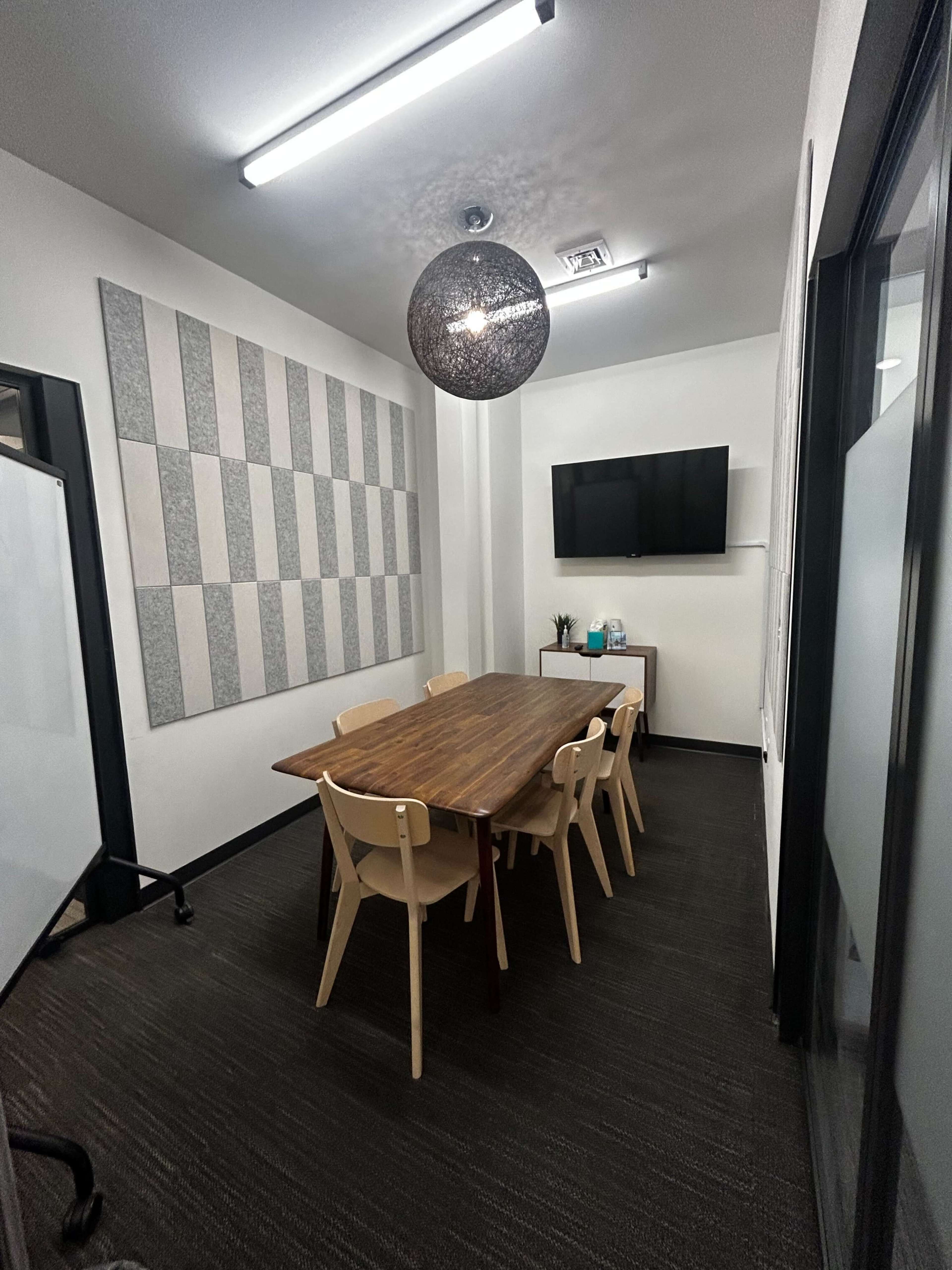 A conference room with a large wooden table surrounded by six chairs, a television mounted on the wall, and acoustic panels on one side.