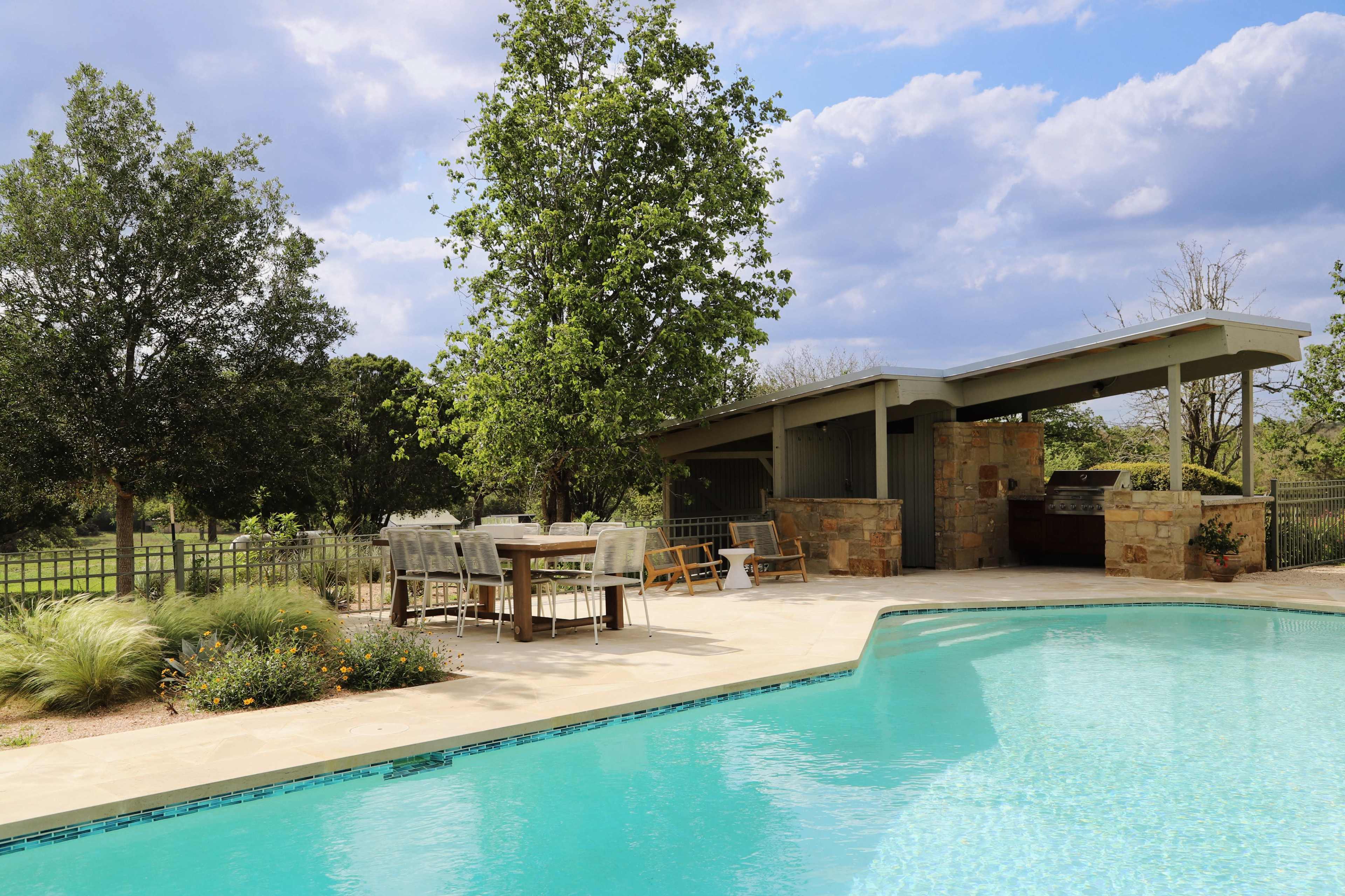 A clear blue swimming pool is surrounded by a patio area with outdoor furniture, a stone structure, and greenery in the background.