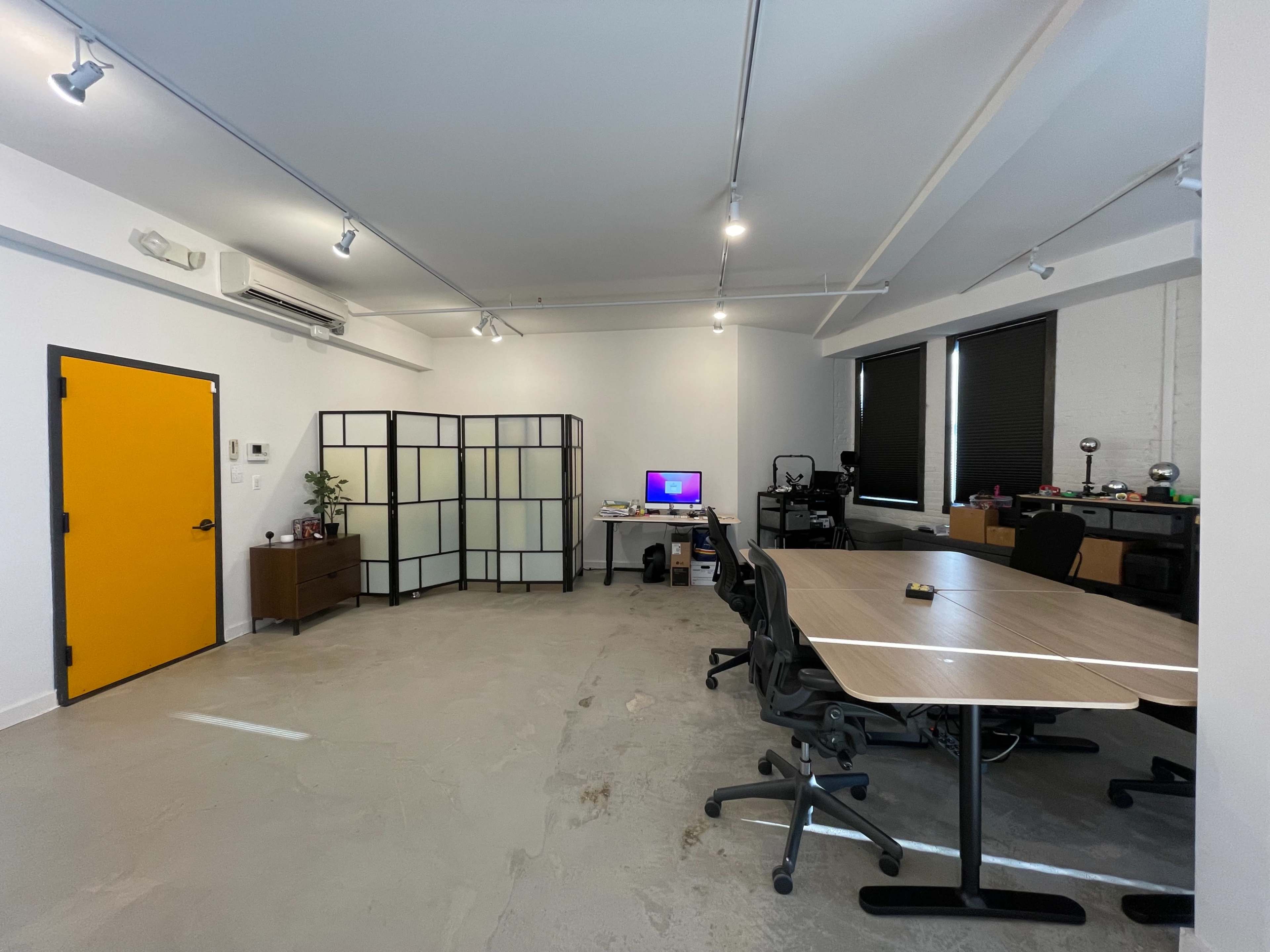 A spacious office featuring a large table with chairs, a partition screen, a computer setup, and a bright yellow door.