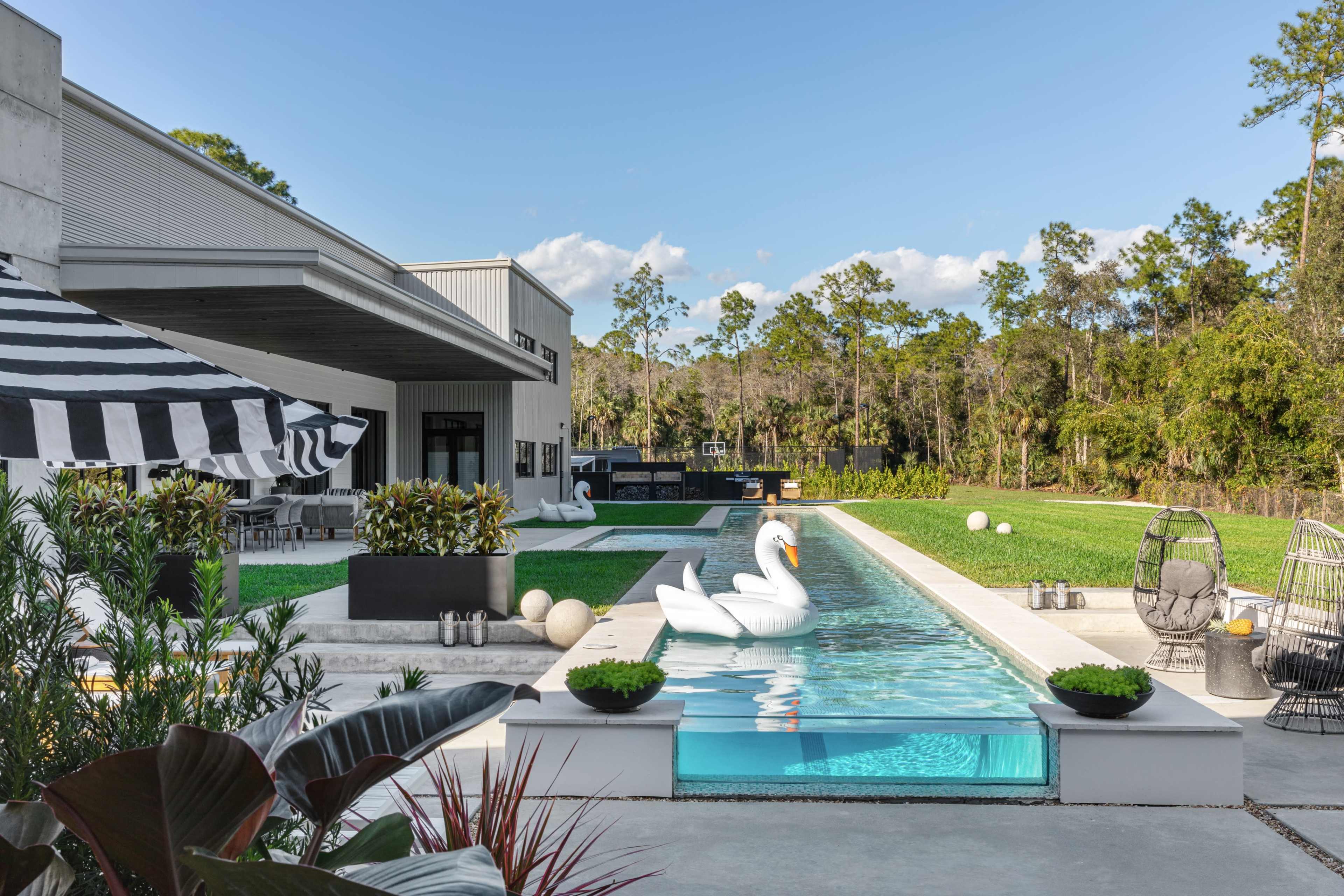A modern backyard features a rectangular swimming pool with a swan float, surrounded by lush green grass, lounge chairs, and patio furniture under a striped awning.