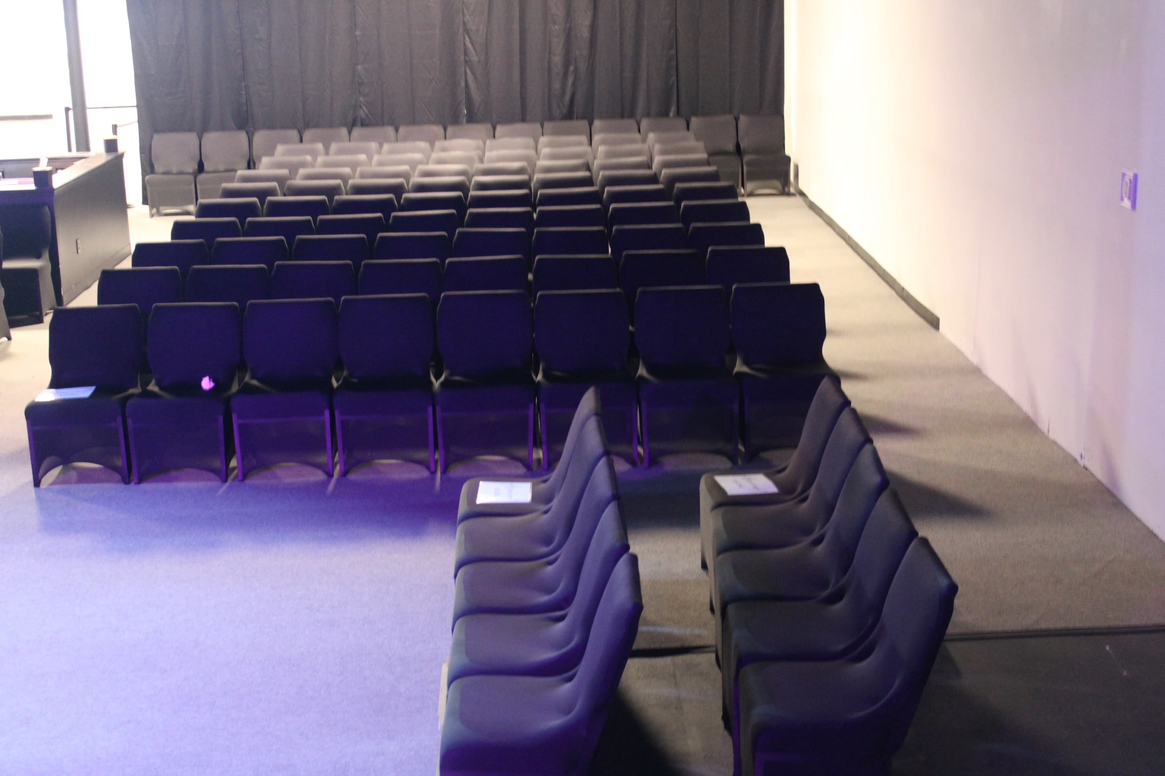 The image shows a seated auditorium with rows of black chairs arranged in a neat, orderly fashion facing a dark backdrop.
