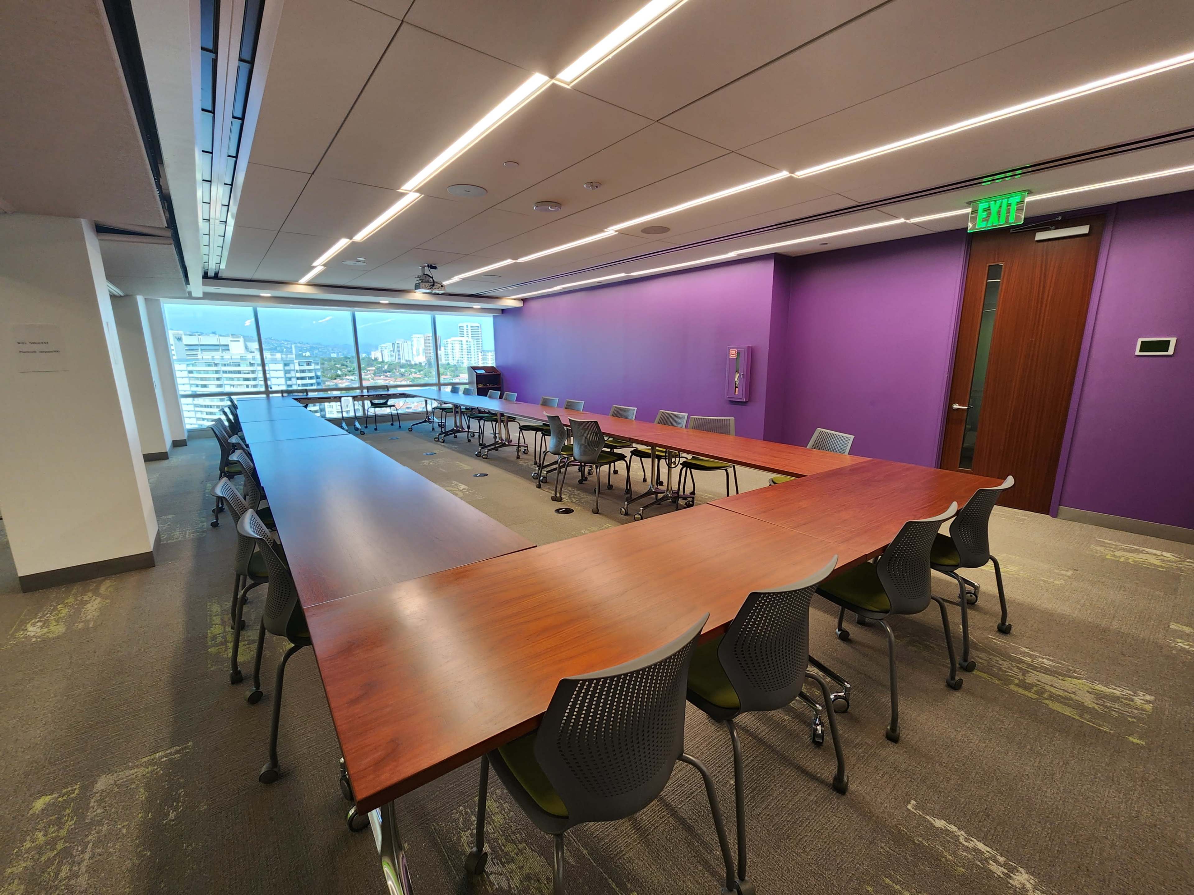 A modern conference room features a large rectangular table surrounded by ergonomic chairs, with a purple accent wall and large windows offering a view of the outside.
