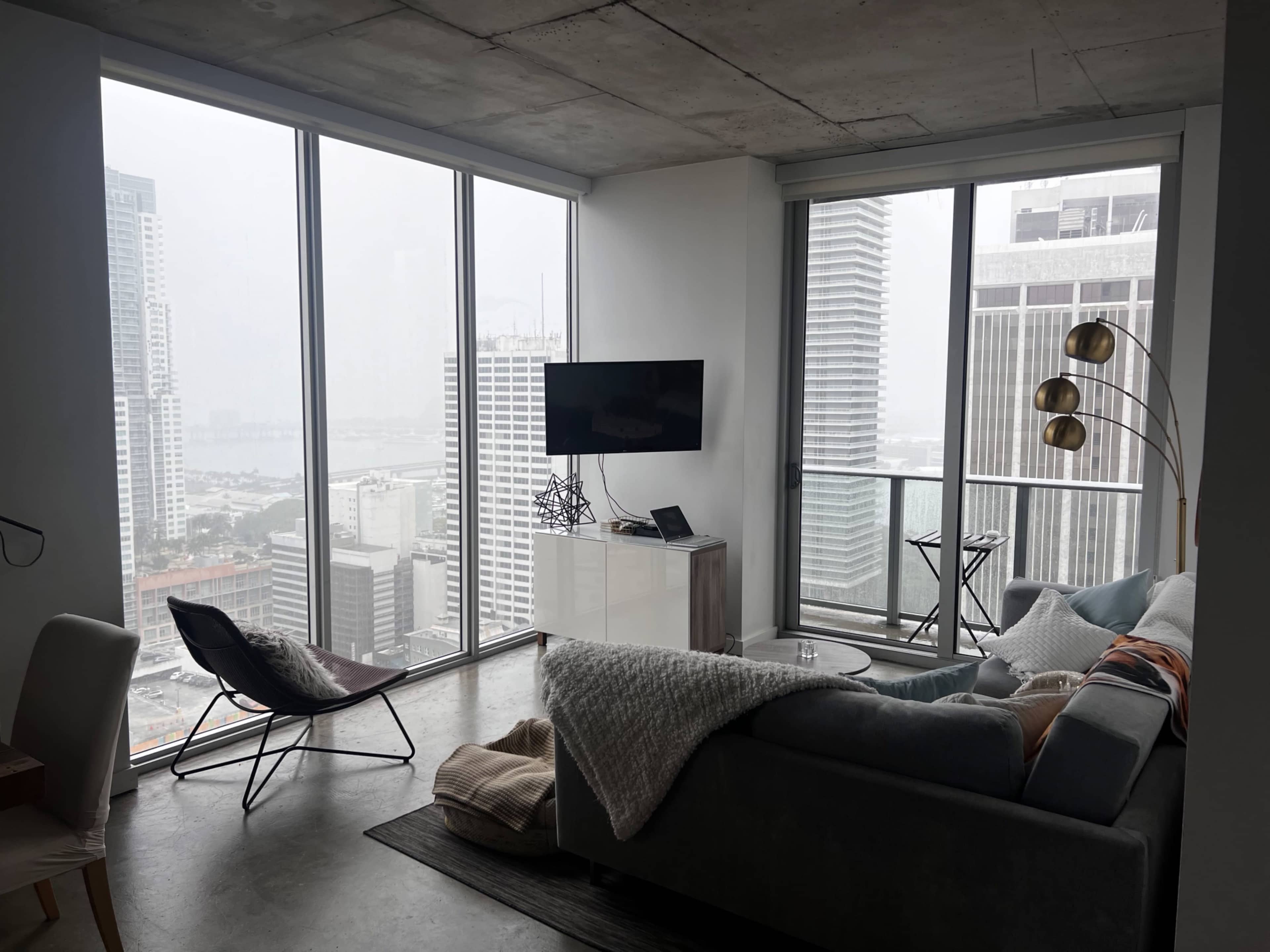 A modern apartment features large windows overlooking a cityscape, with a cozy living area that includes a sofa, television, and a floor lamp.