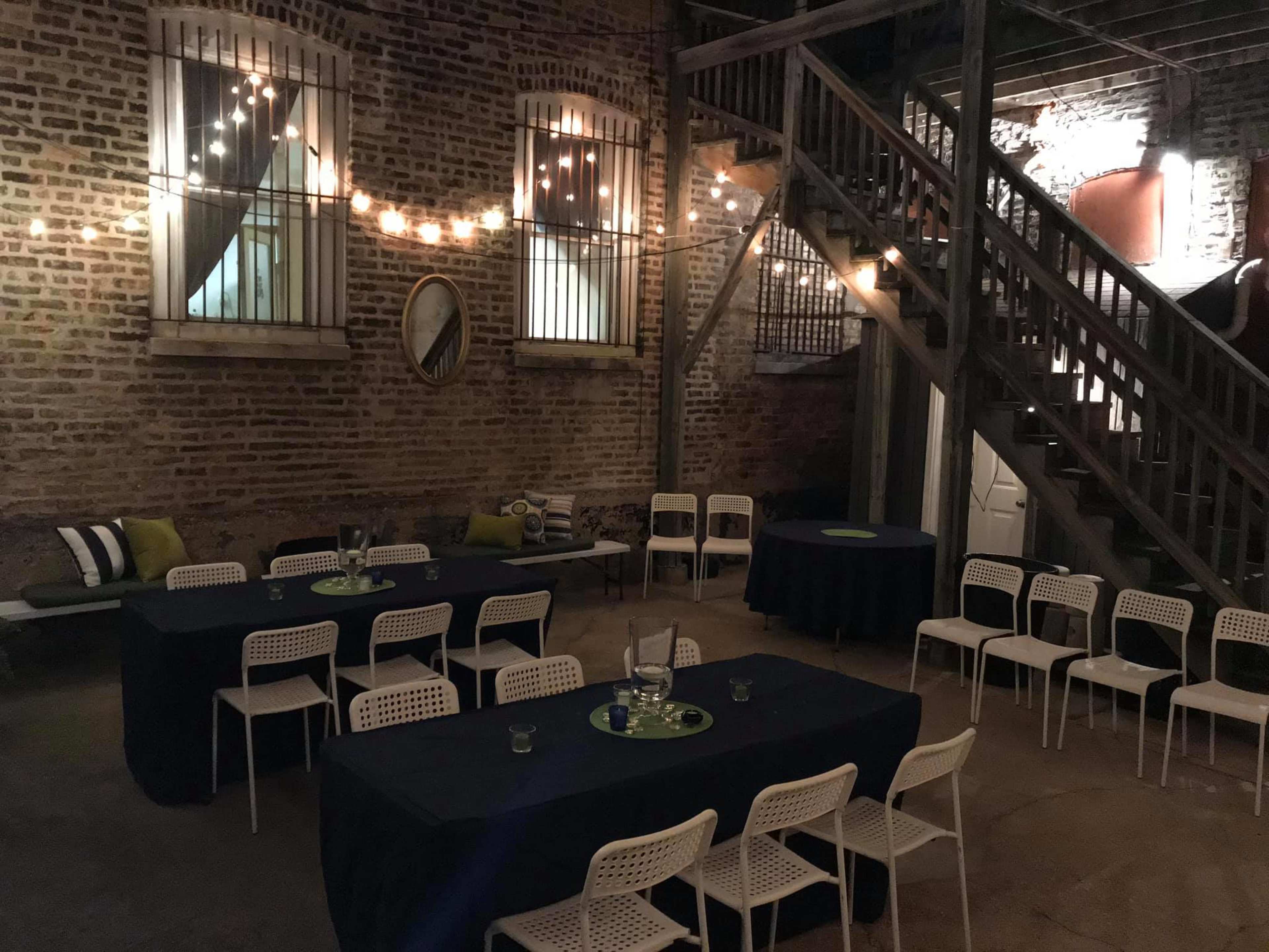 The image shows an indoor event space with brick walls, string lights, tables covered in dark cloth, and white chairs arranged around them.