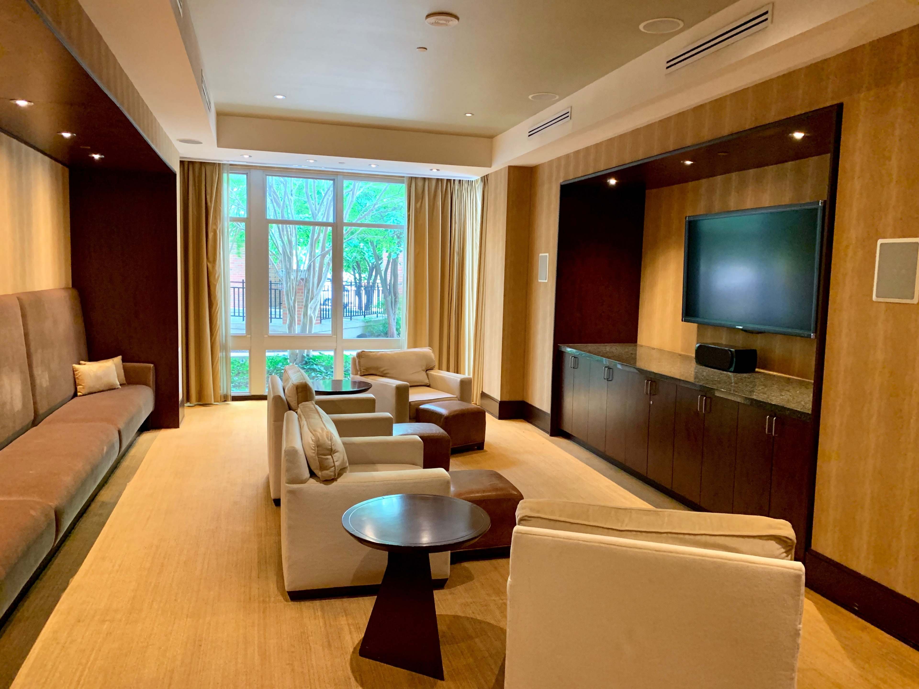 A modern lounge area with a sofa, chairs, a coffee table, and a television against a wall, illuminated by natural light coming from large windows.