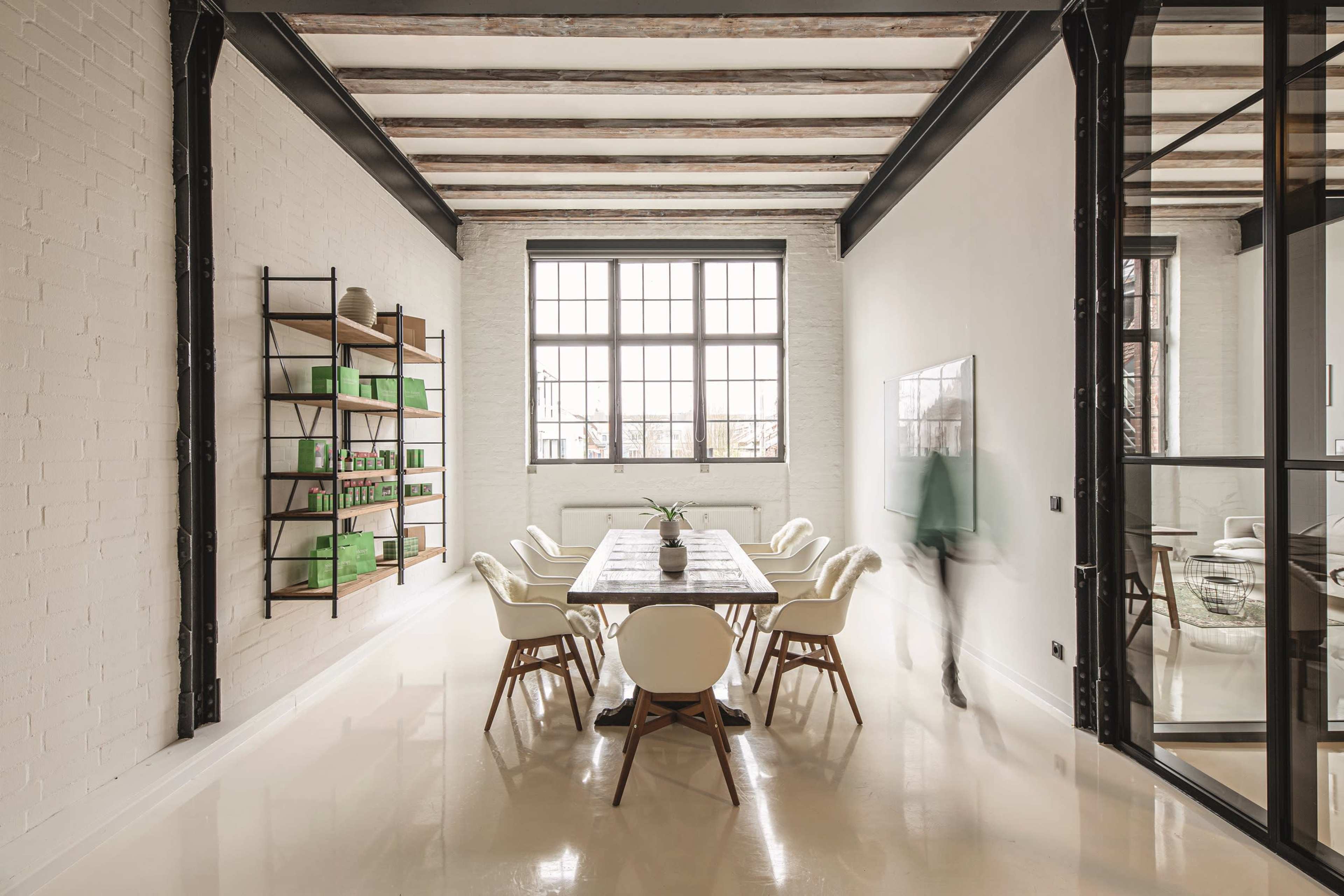 A long dining table with white chairs is set in a spacious room featuring large windows and exposed wooden beams.