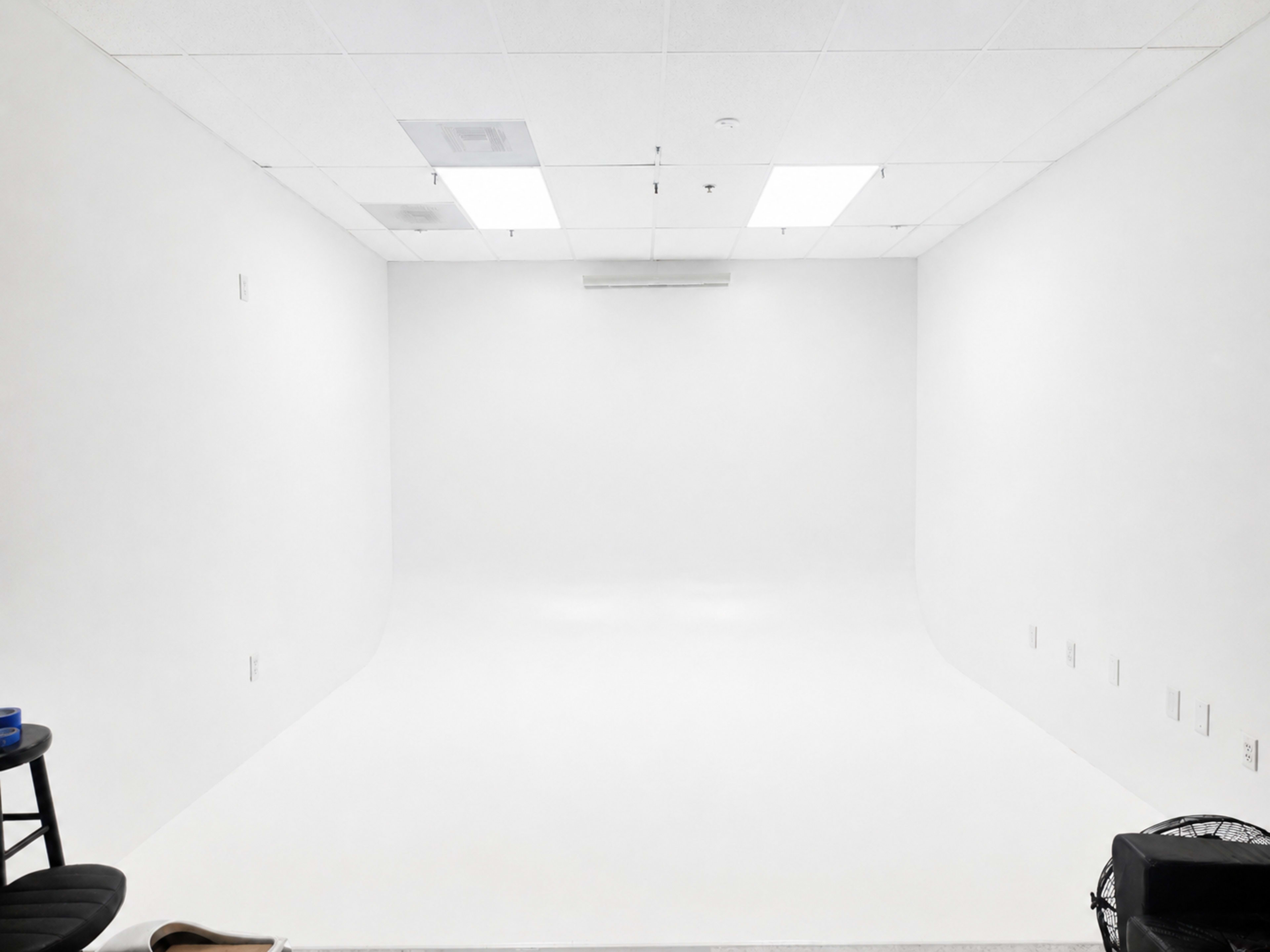 The image shows a spacious, empty room with white walls, a seamless white floor, and bright overhead lighting.