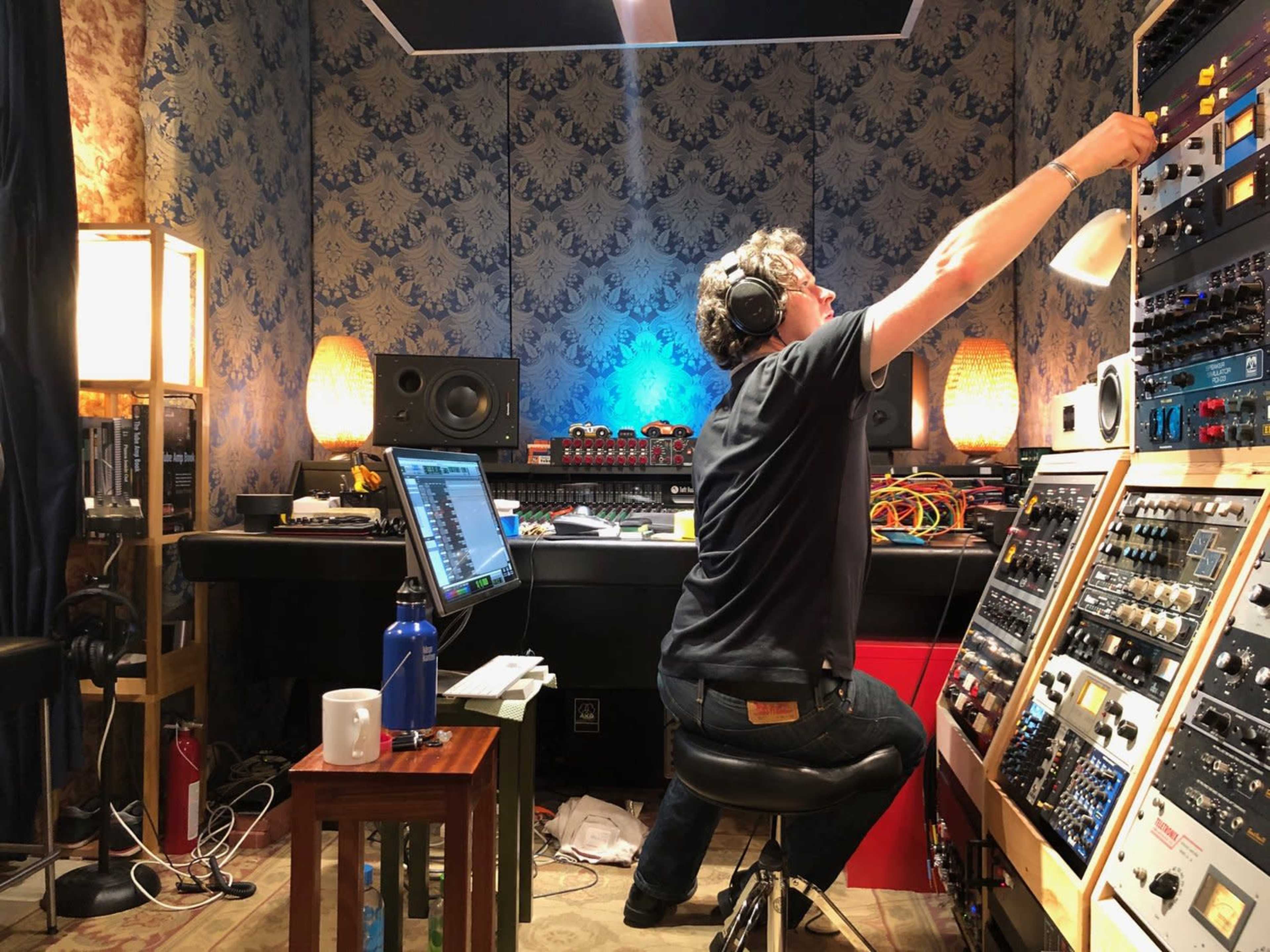 A person adjusts equipment on a sound mixer in a dimly lit music studio filled with various instruments and recording gear.