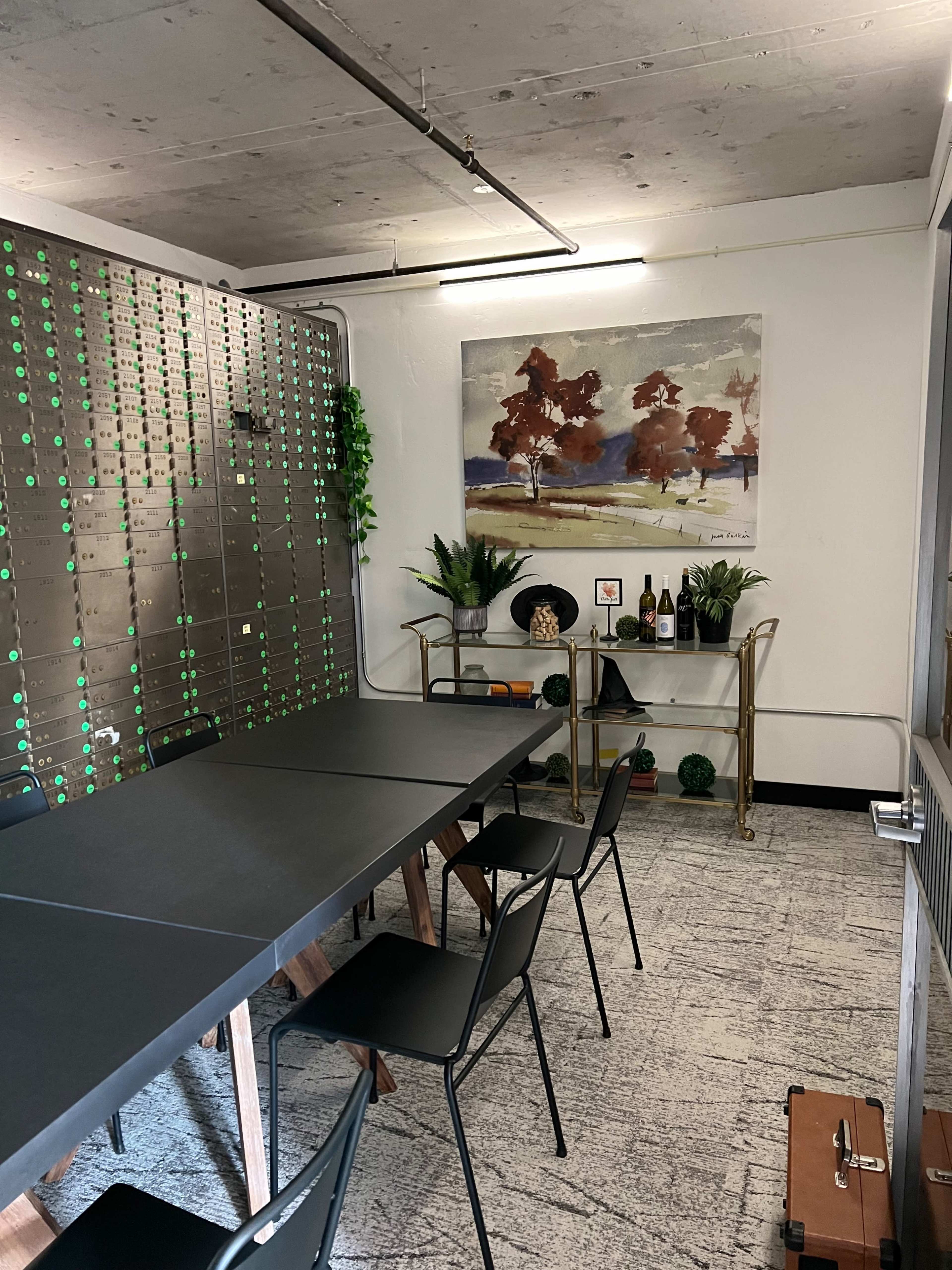 A modern meeting room with a long black table, black chairs, a large framed painting on the wall, and a side table displaying plants and bottles.
