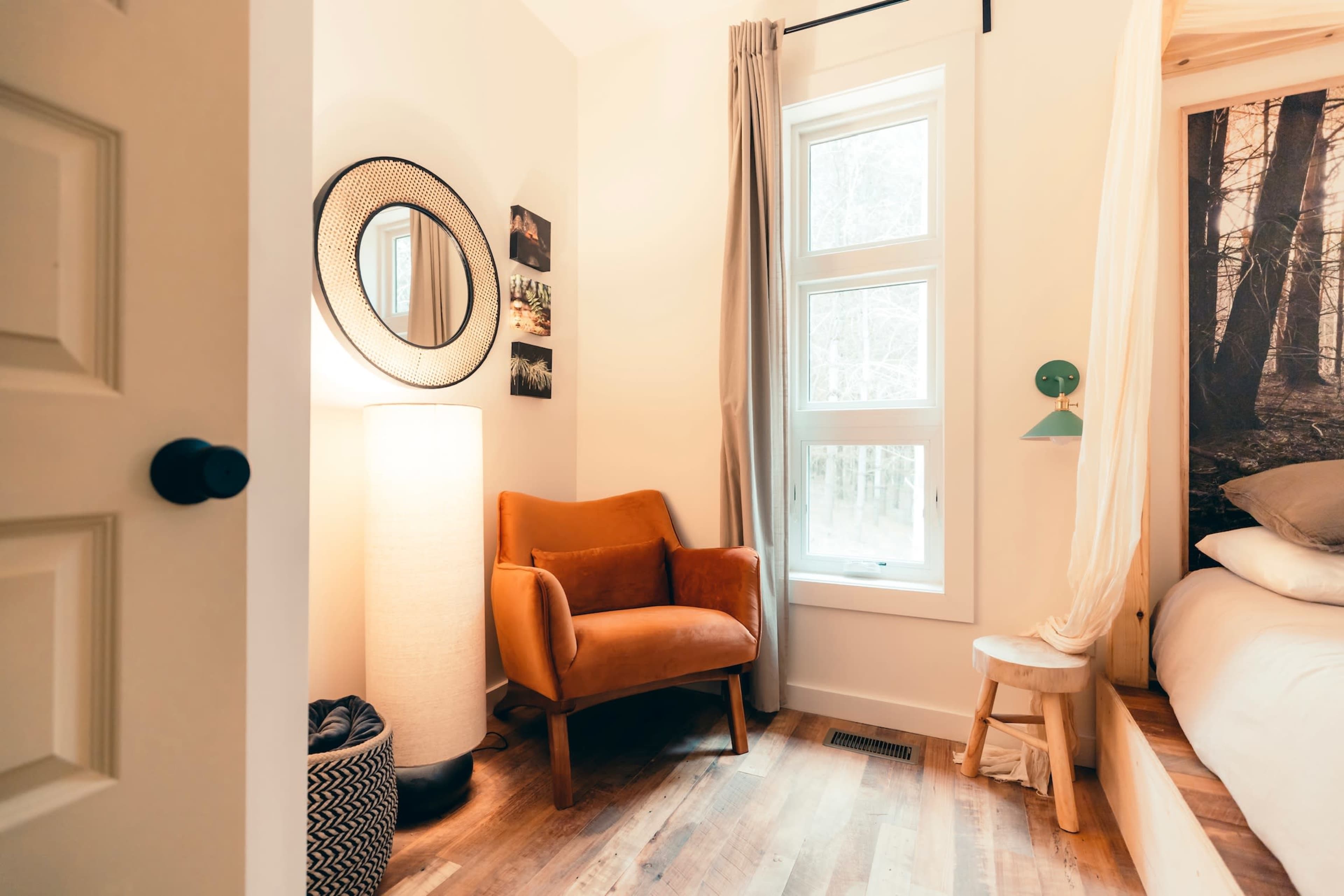 A cozy bedroom featuring a brown armchair, a tall lamp, and a window with light curtains, along with natural wood flooring.