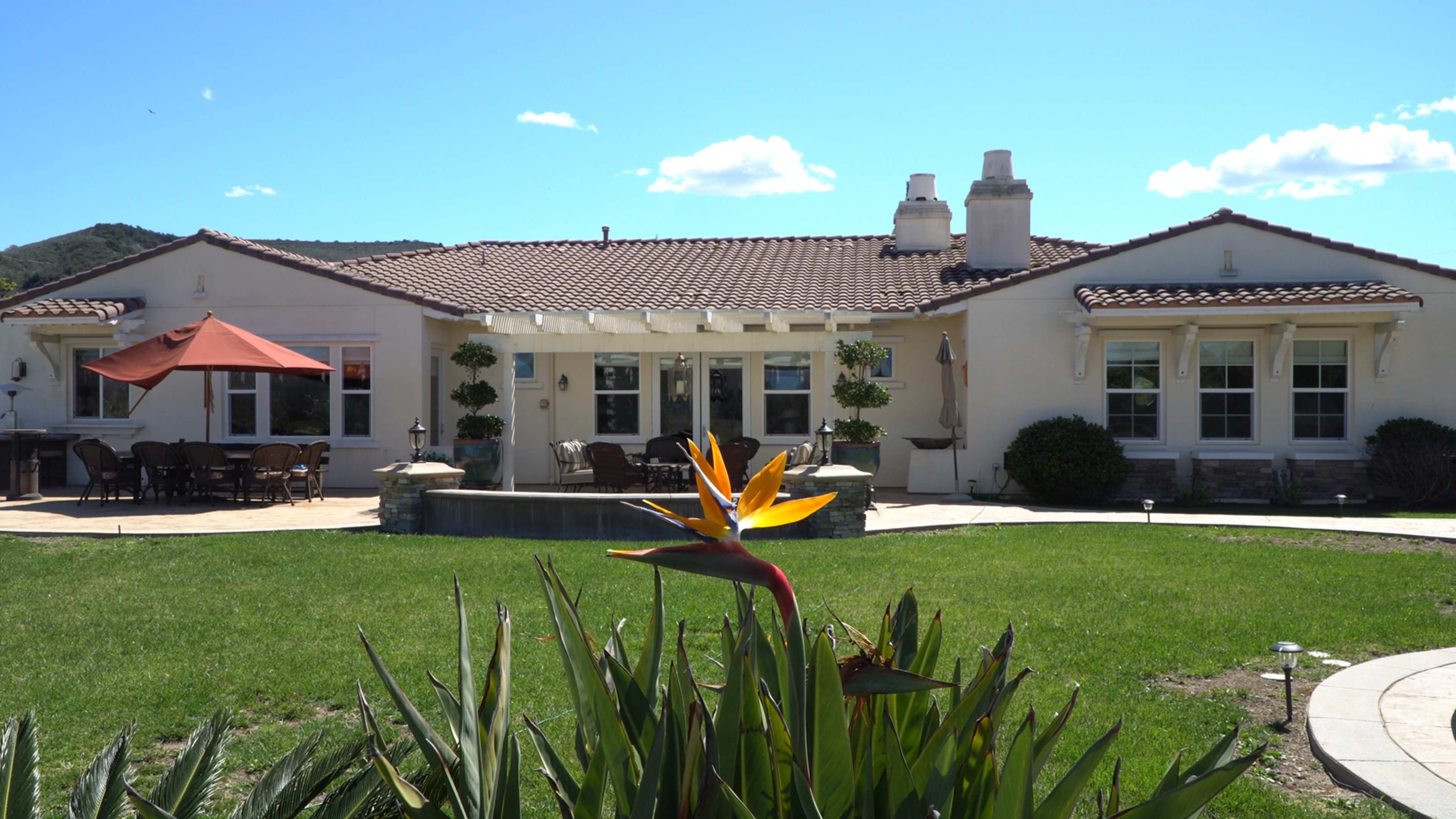 A single-story house with a tiled roof, outdoor seating area, and landscaped garden is set against a clear blue sky.