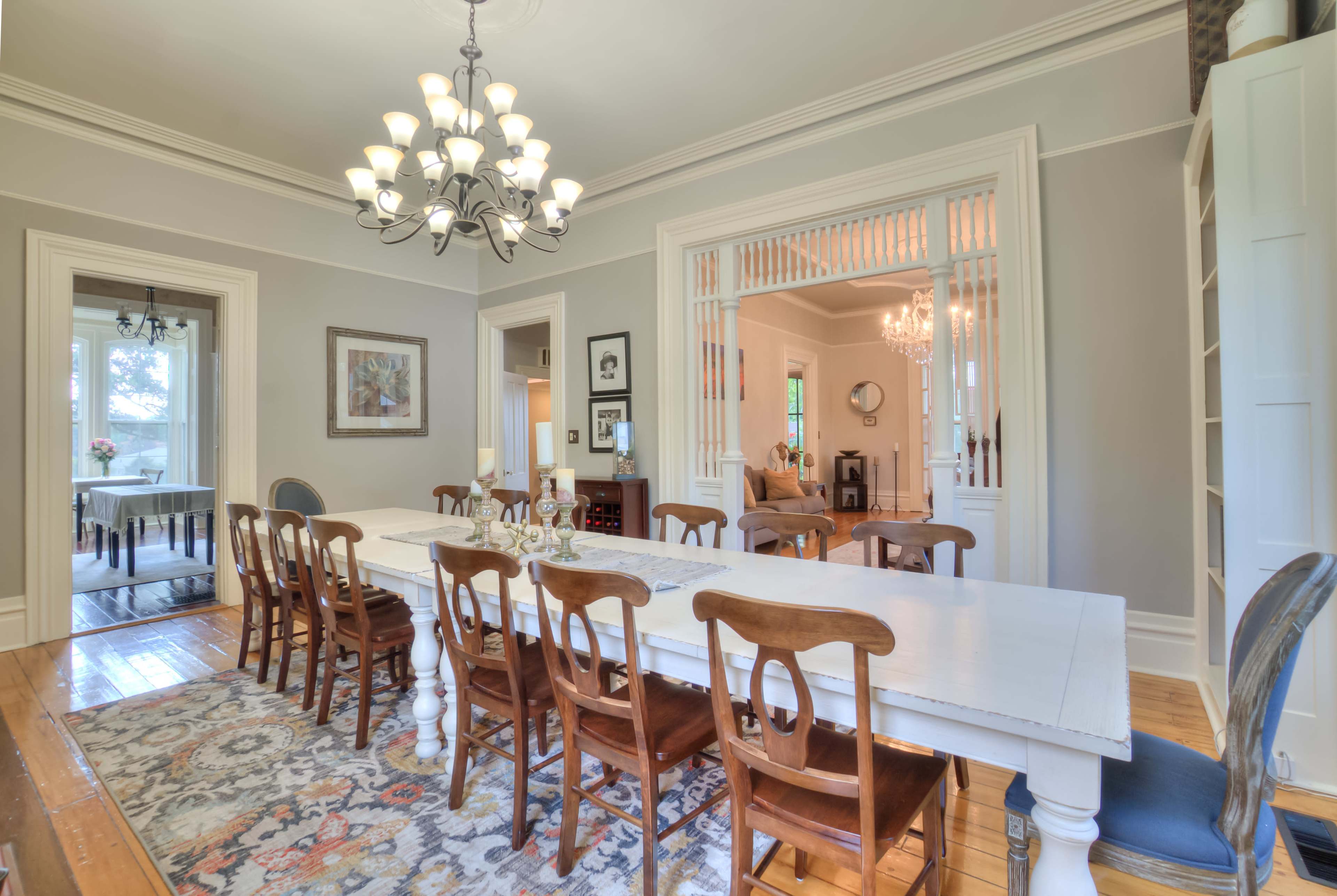 A spacious dining room with a long, white table surrounded by wooden chairs, illuminated by a chandelier, with an adjoining area visible through a decorative partition.