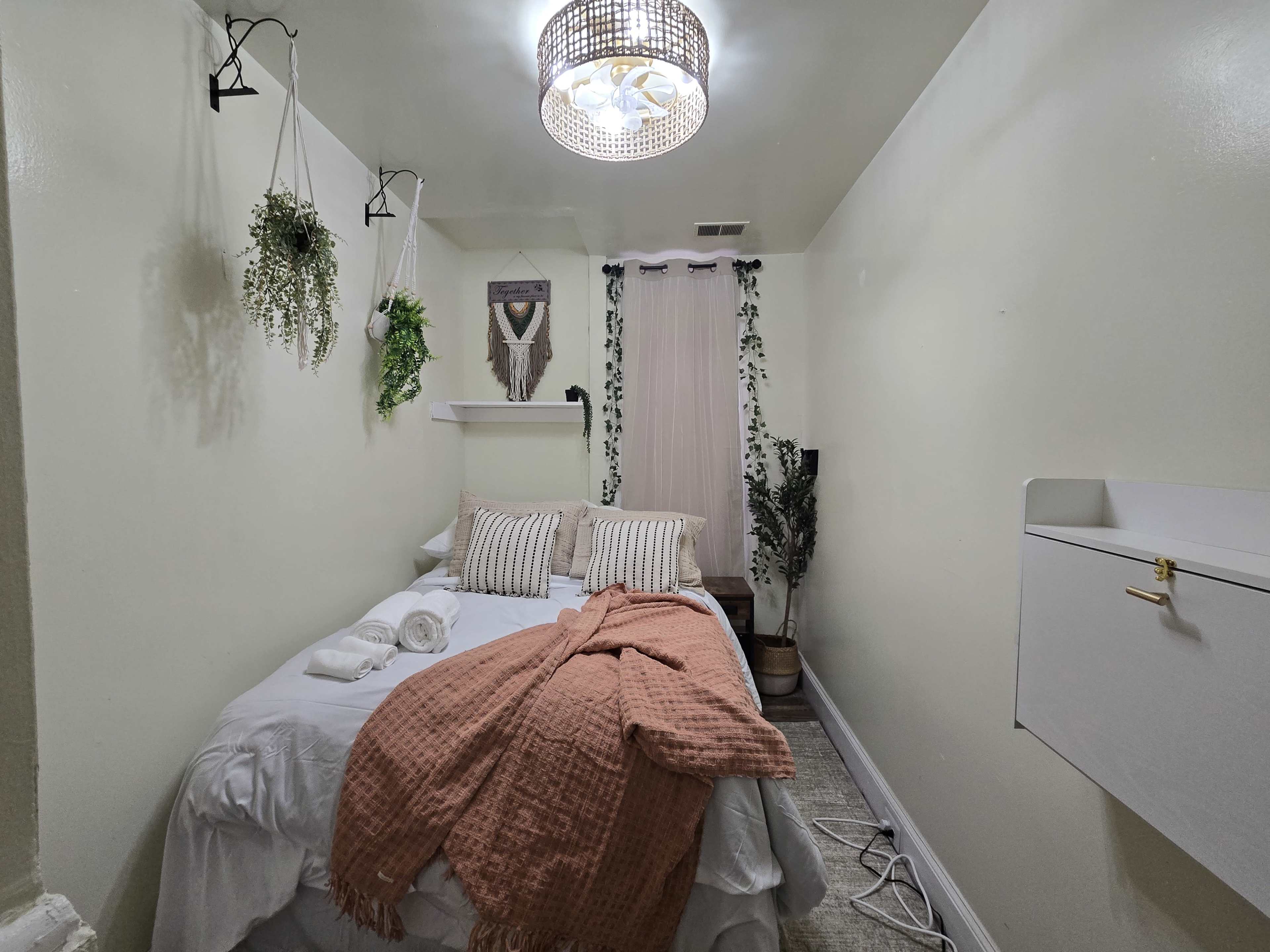 The image shows a small, cozy bedroom with a bed adorned with a pink blanket, decorative pillows, and plants hanging on the walls.