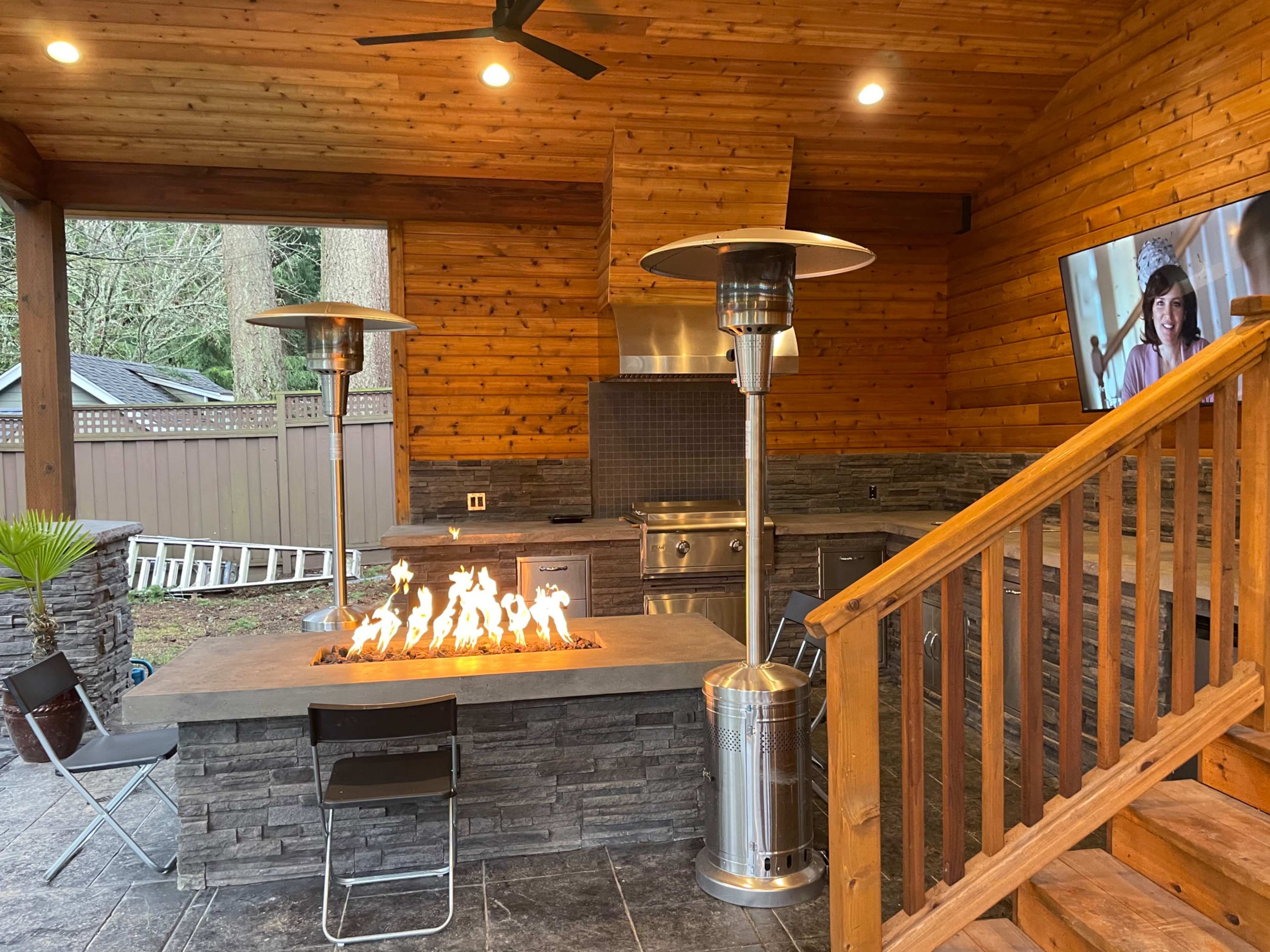 An outdoor kitchen area with a stone fire pit, metal patio heaters, and a television mounted on the wooden wall.