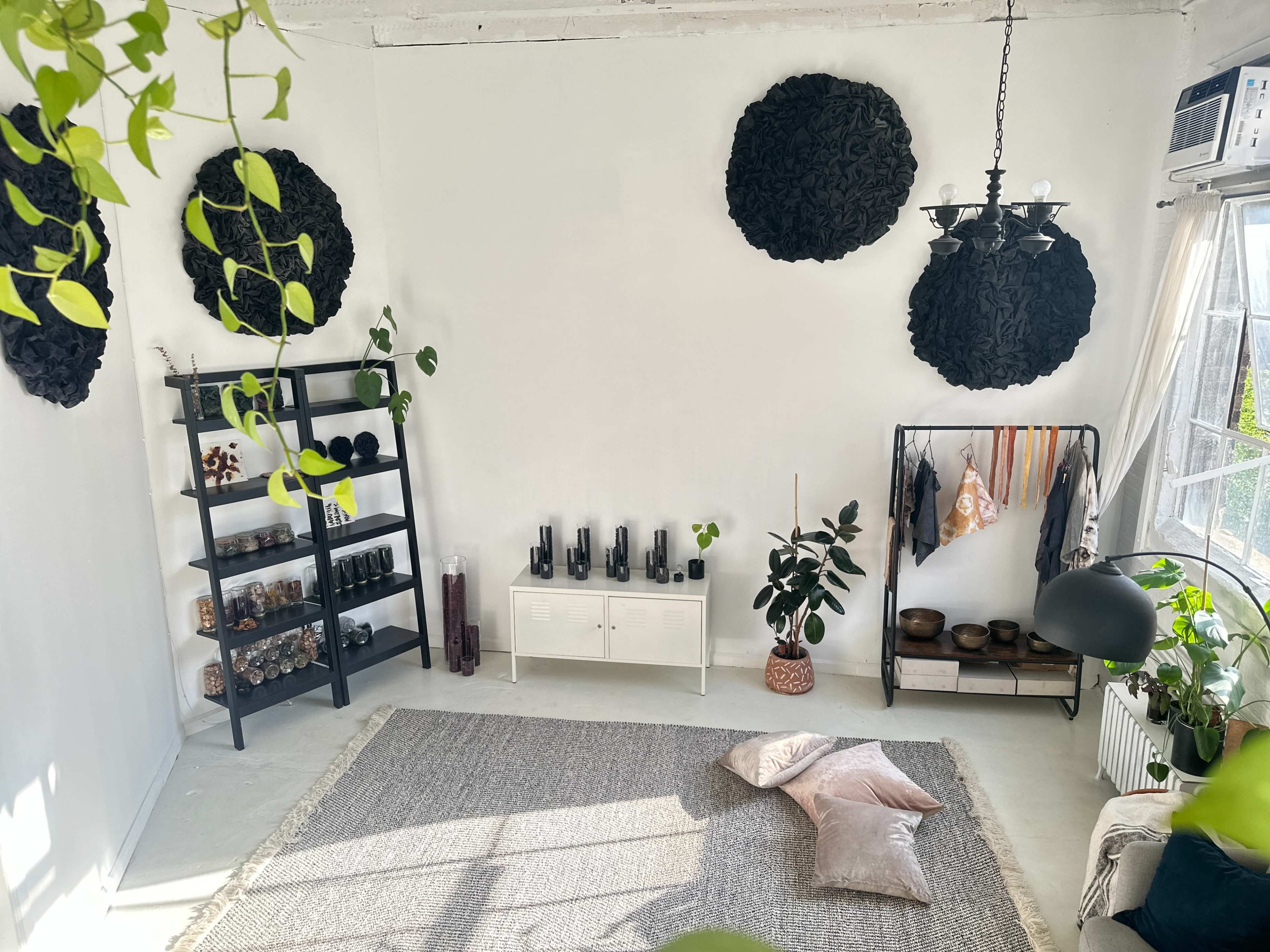 The image shows a minimalist interior space with black wall decorations, a shelving unit filled with jars and glassware, a soft rug with pillows on the floor, and plants in various locations.