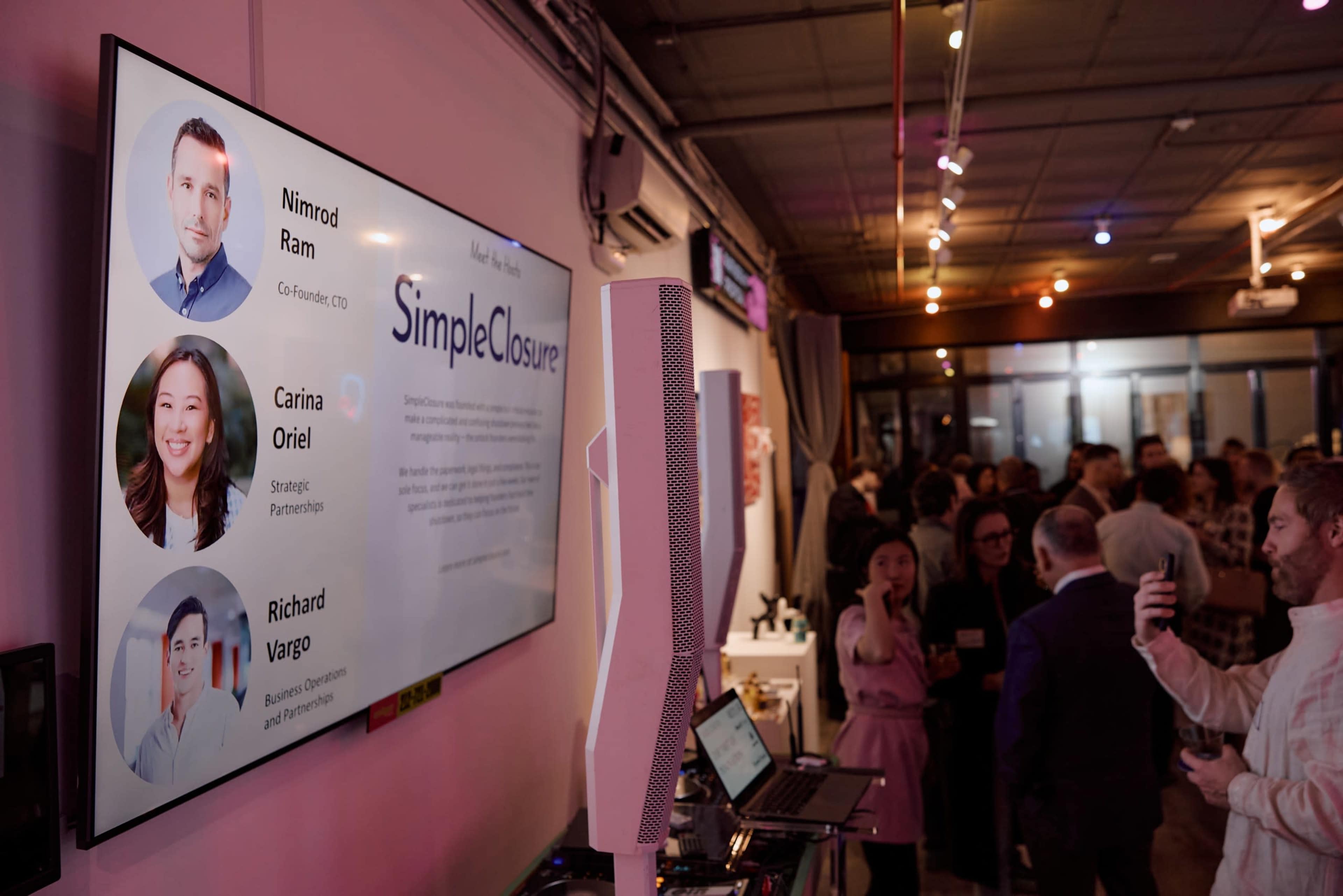 A digital display shows profiles of three individuals associated with SimpleClosure at a networking event, with attendees mingling in the background.