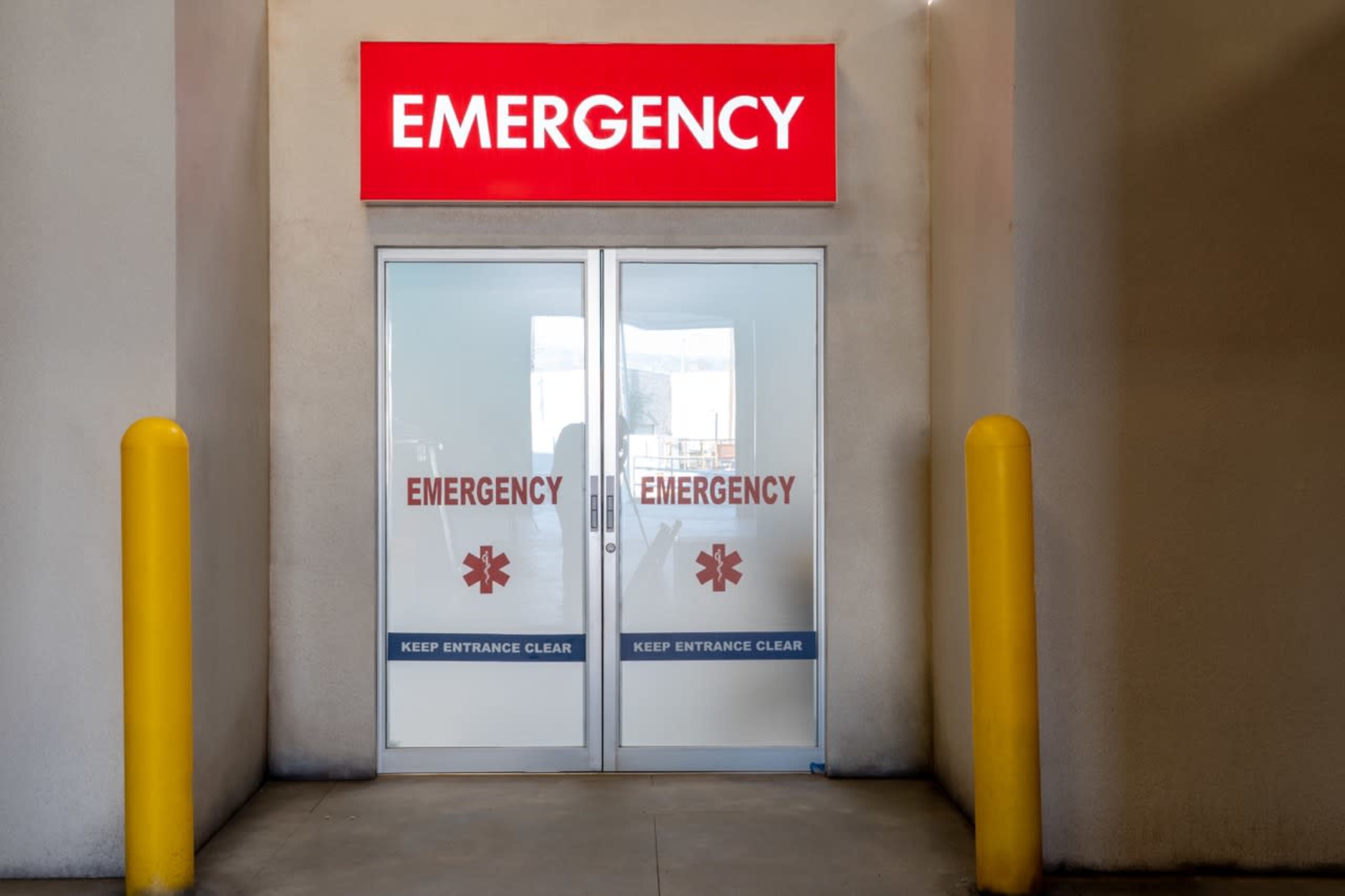 Double glass doors marked with the word "EMERGENCY" in bold red letters, flanked by yellow bollards.