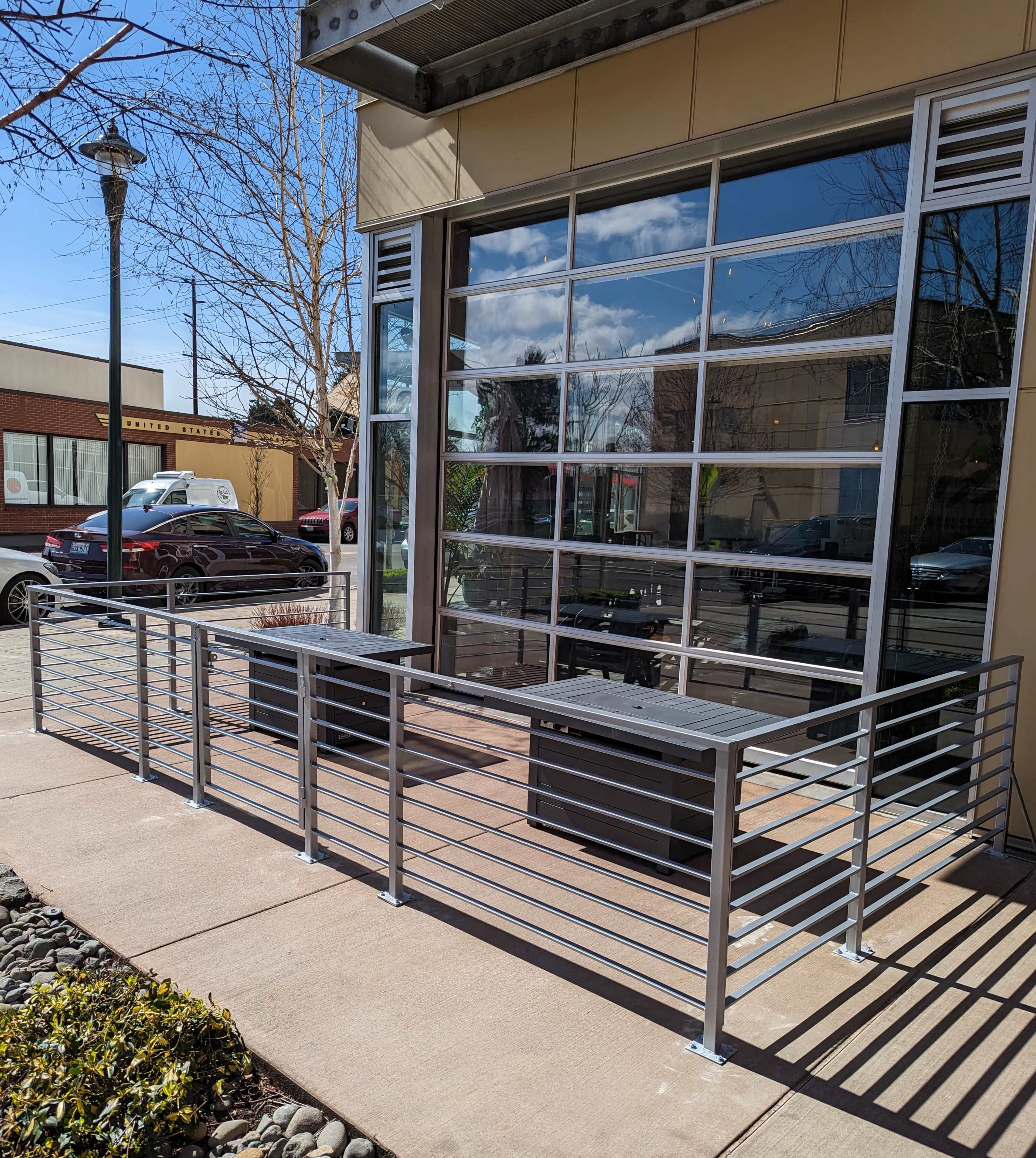 A metal railing enclosing a small outdoor seating area in front of a building with large glass windows.